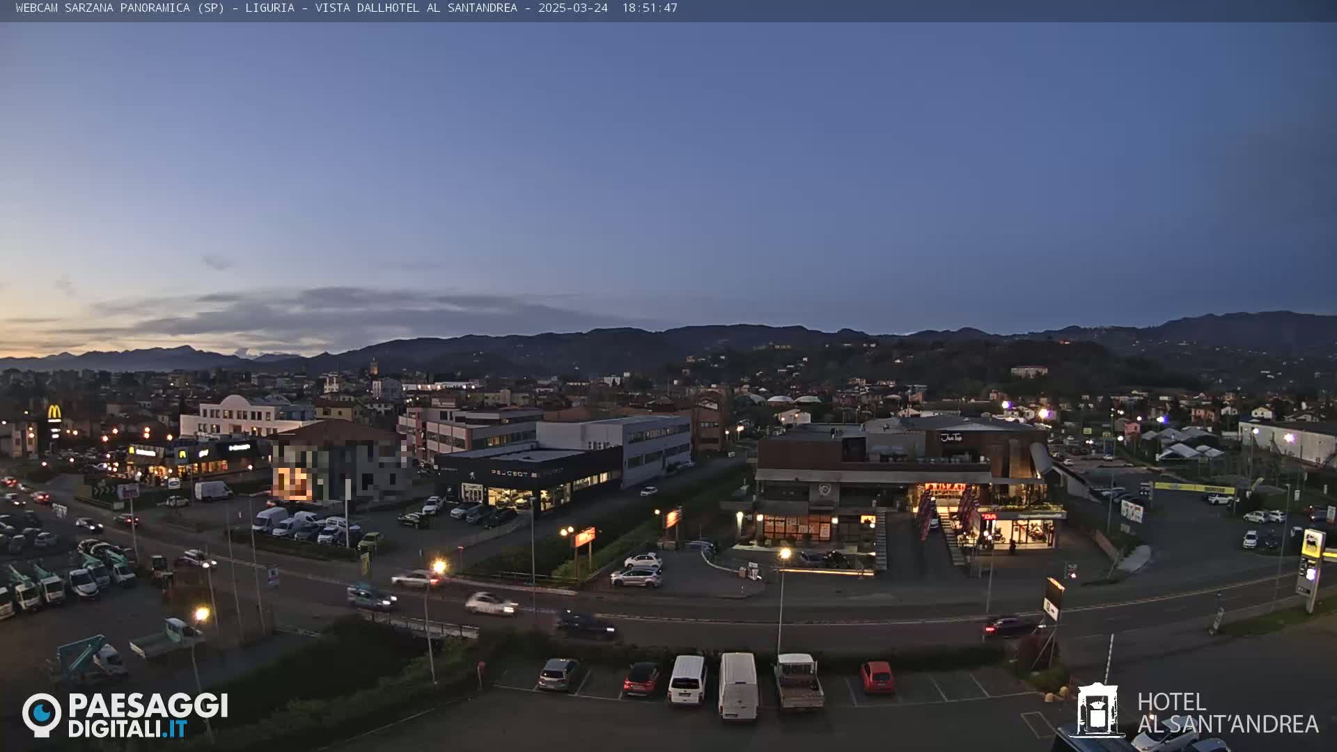 A twilight view of a town with mountains in the background, featuring streets, buildings, parking lots, and cars, under a clear, dark blue sky.
