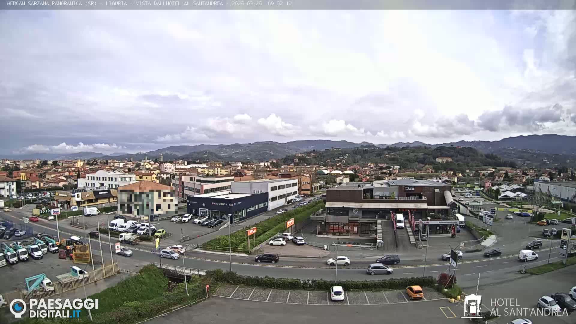 A mostly cloudy day reveals a sprawling town nestled in a valley surrounded by low mountains, with numerous cars parked in lots surrounding various buildings.