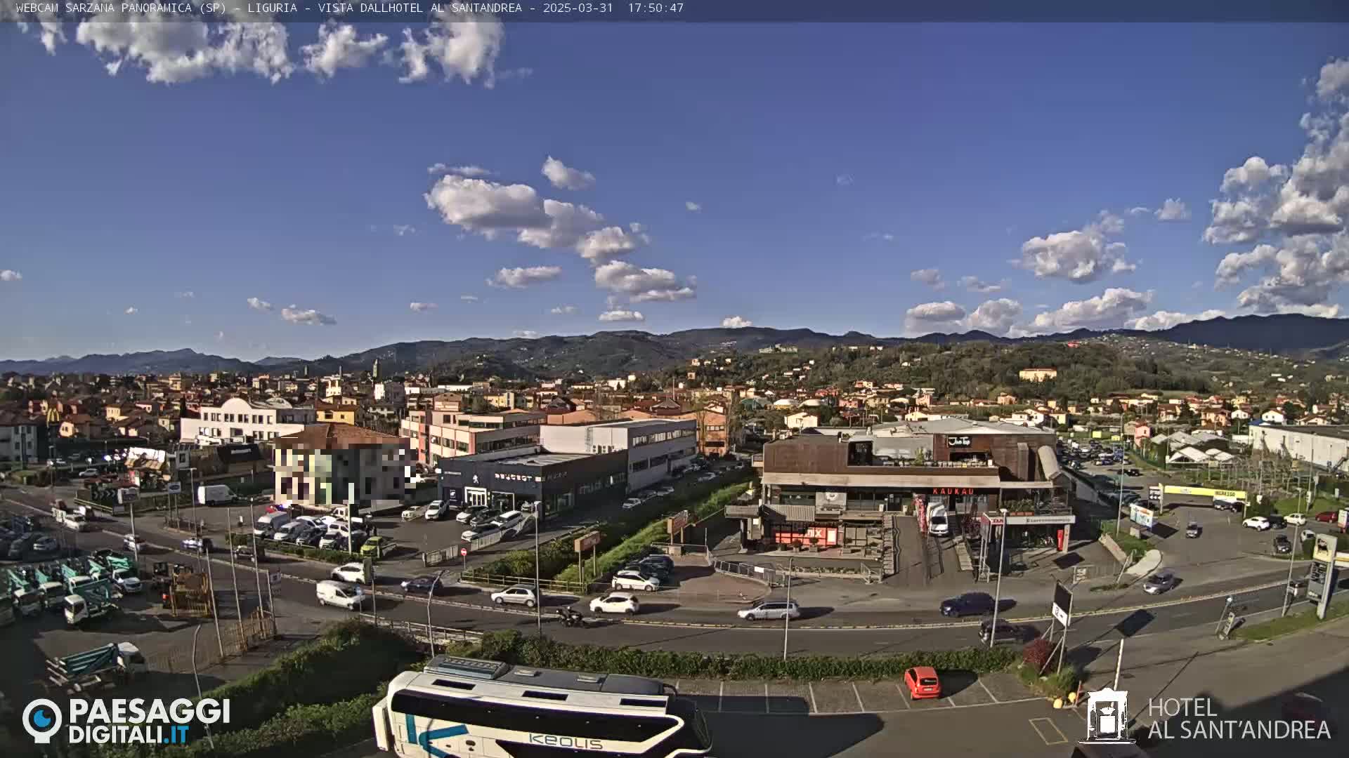 A sunny day with scattered clouds reveals a wide shot of a town situated in a valley, surrounded by hills, with cars parked in a parking lot and a large bus visible in the foreground.
