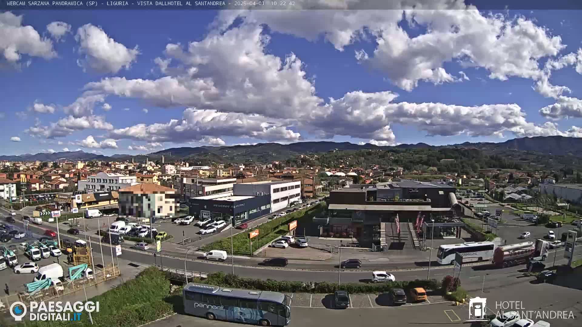 A partly cloudy day reveals a panoramic view of a town with numerous buildings, cars, and buses in parking lots and on roadways, nestled at the foot of distant hills.