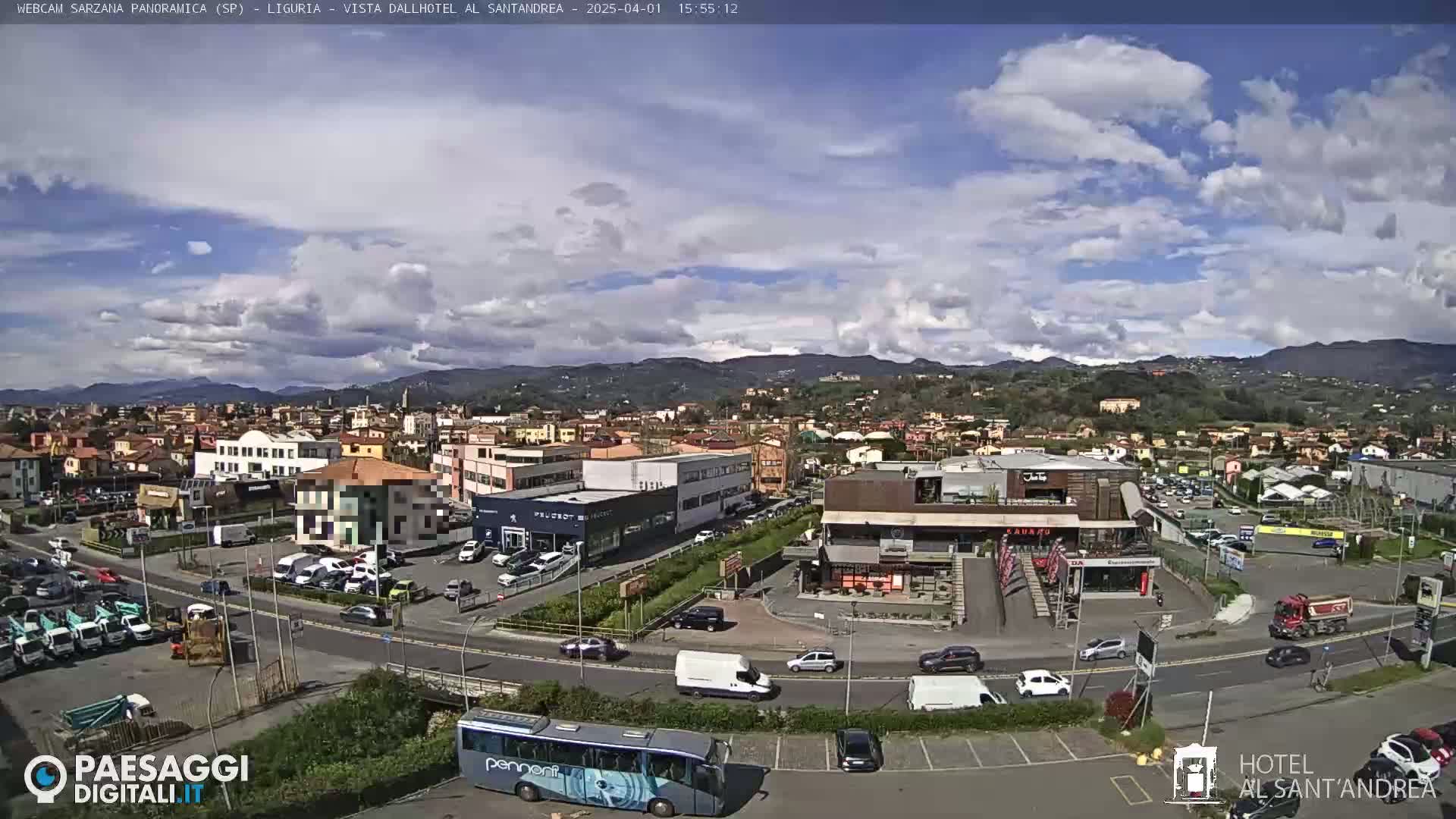 A partly cloudy day reveals a view of a town with buildings, roads, cars, and a bus, nestled among hills.