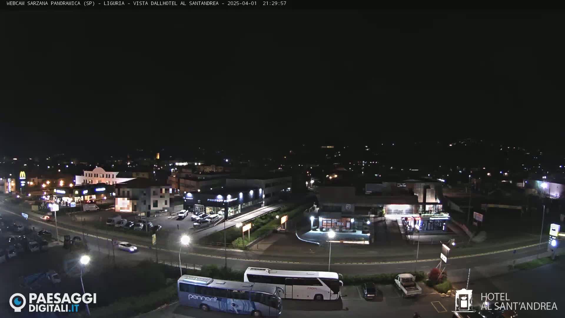 A nighttime aerial view of a town's illuminated streets and buildings, including a parking lot with several cars and two large buses, under a clear, dark sky.
