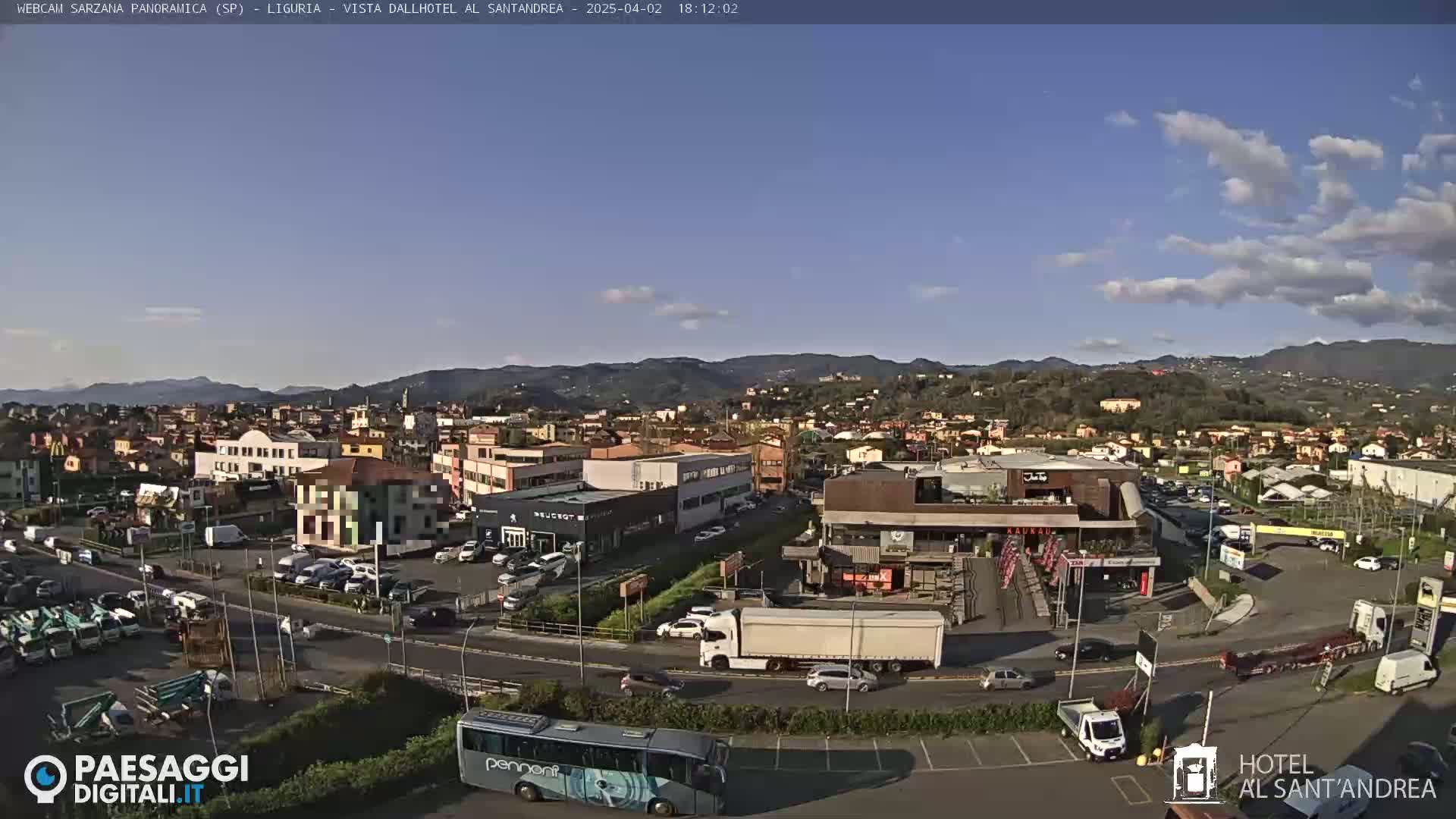 A sunny day reveals a panoramic view of a town nestled in a valley, with mountains in the background and various vehicles including buses and trucks visible in a parking lot and on roads.