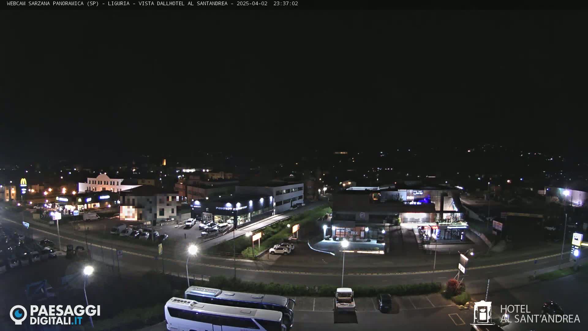 A nighttime, overhead view of a town's streets and buildings, including a parking lot with a bus and cars, and various businesses with illuminated signs.