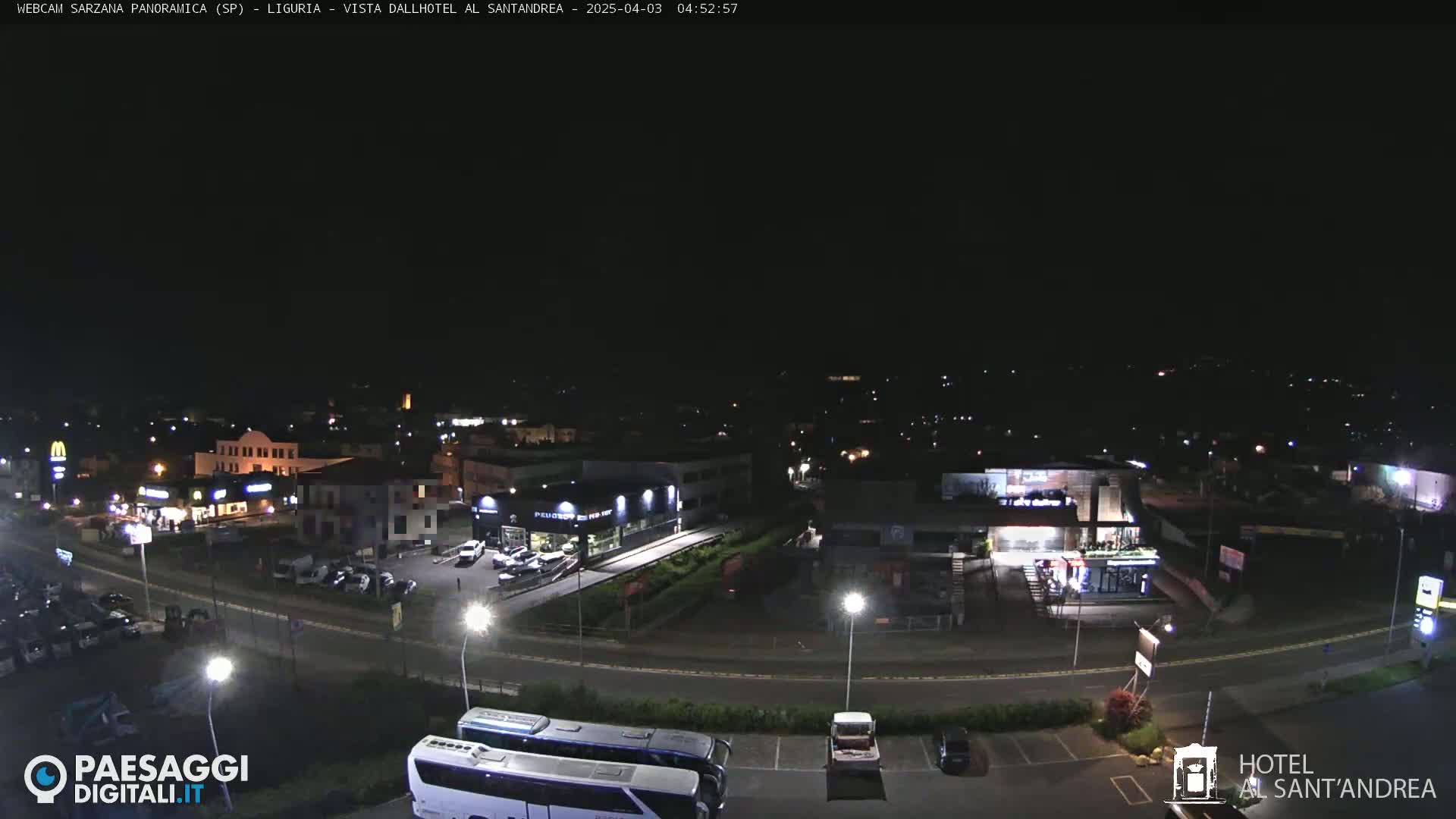 A nighttime, aerial view of a town's outskirts, showing roadways, buildings with illuminated signage, and parked vehicles.