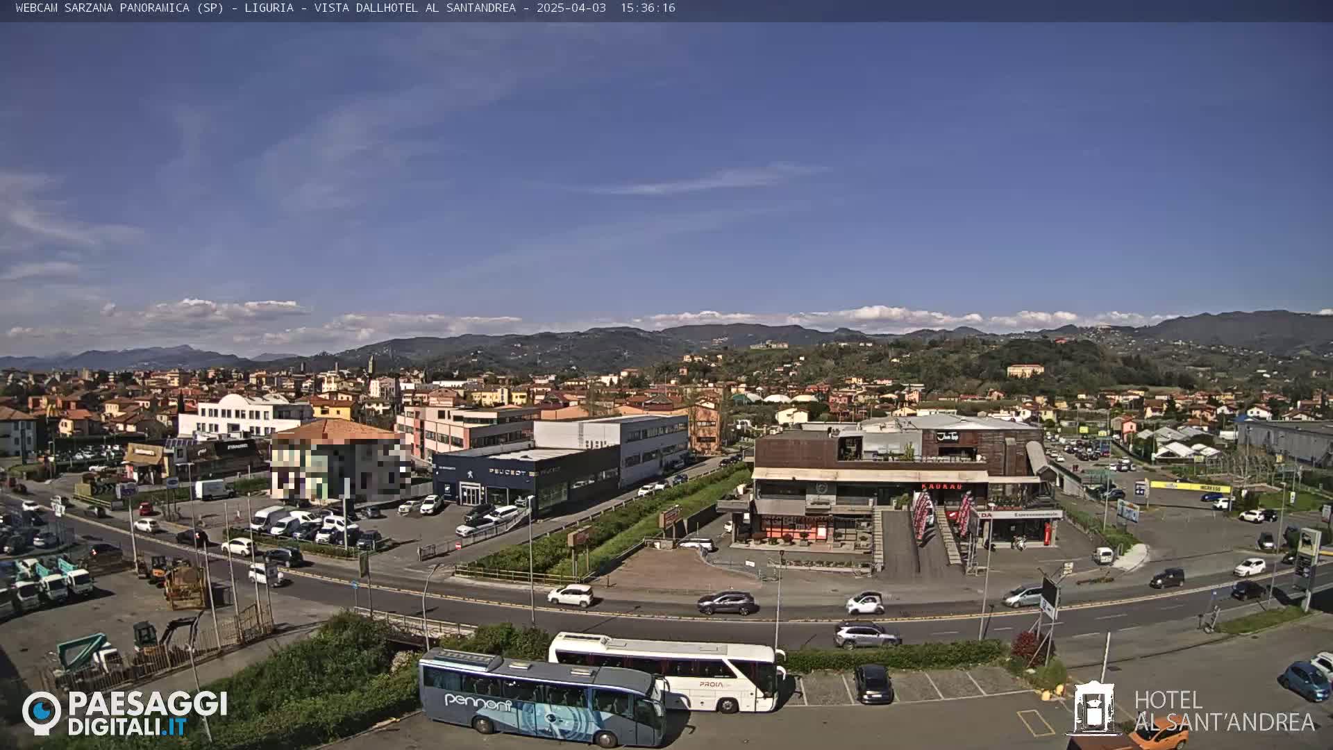 A sunny day reveals a panoramic view of a town nestled in a valley, with numerous buildings, vehicles, and two large buses visible in a parking lot near a roadway.