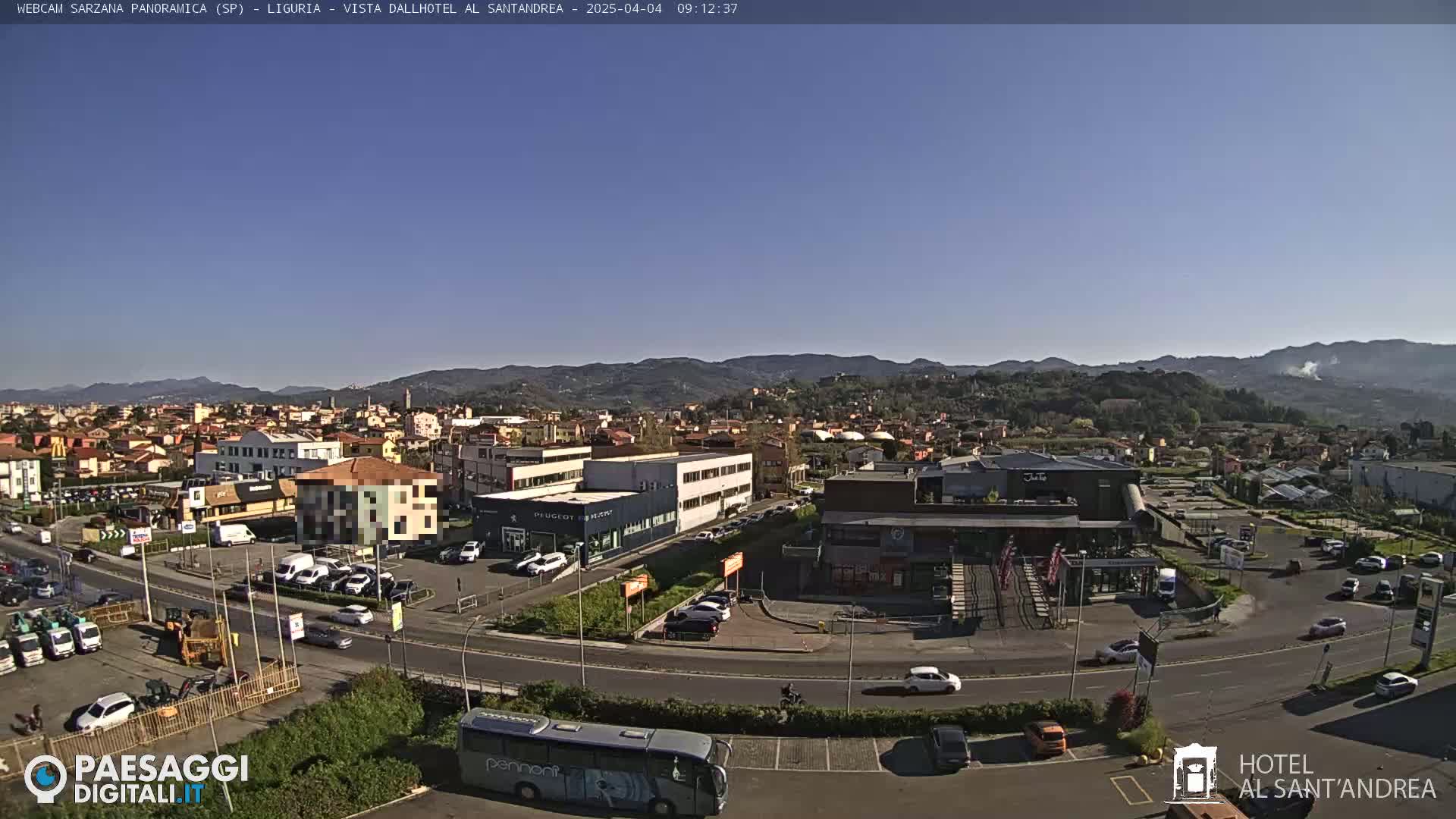 A sunny day reveals a panoramic view of a town nestled among hills, with cars parked in lots and a bus on a road.