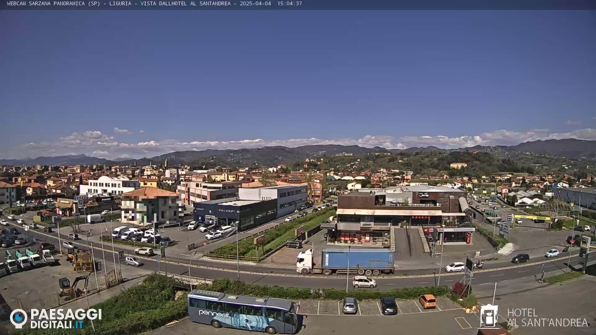 A sunny day reveals a panoramic view of a town nestled among hills, with a parking lot containing cars, buses, and trucks in the foreground.