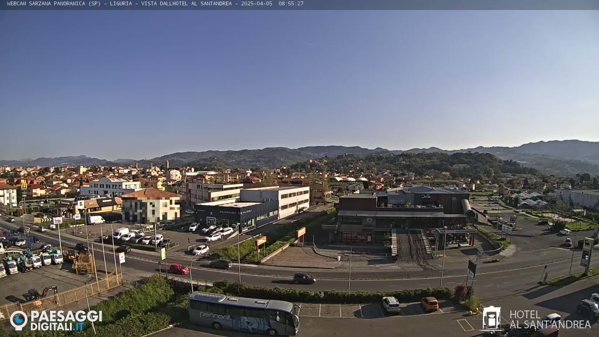 A sunny day reveals a sprawling town nestled against a backdrop of rolling hills, with various buildings, vehicles, and a bus visible.