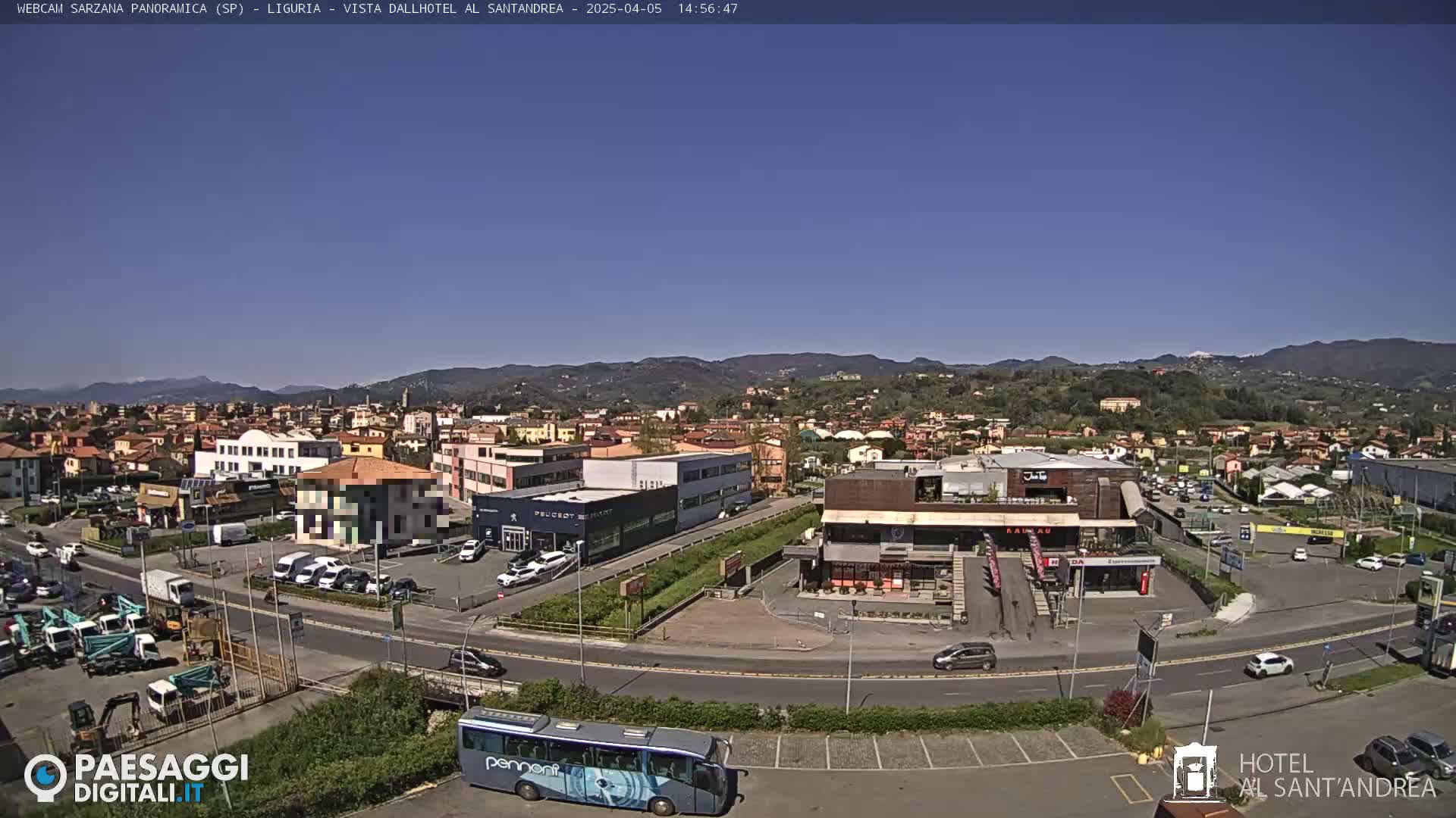 A clear, sunny day reveals a panoramic view of a town, featuring buildings, vehicles, and a large bus, with mountains visible in the background.