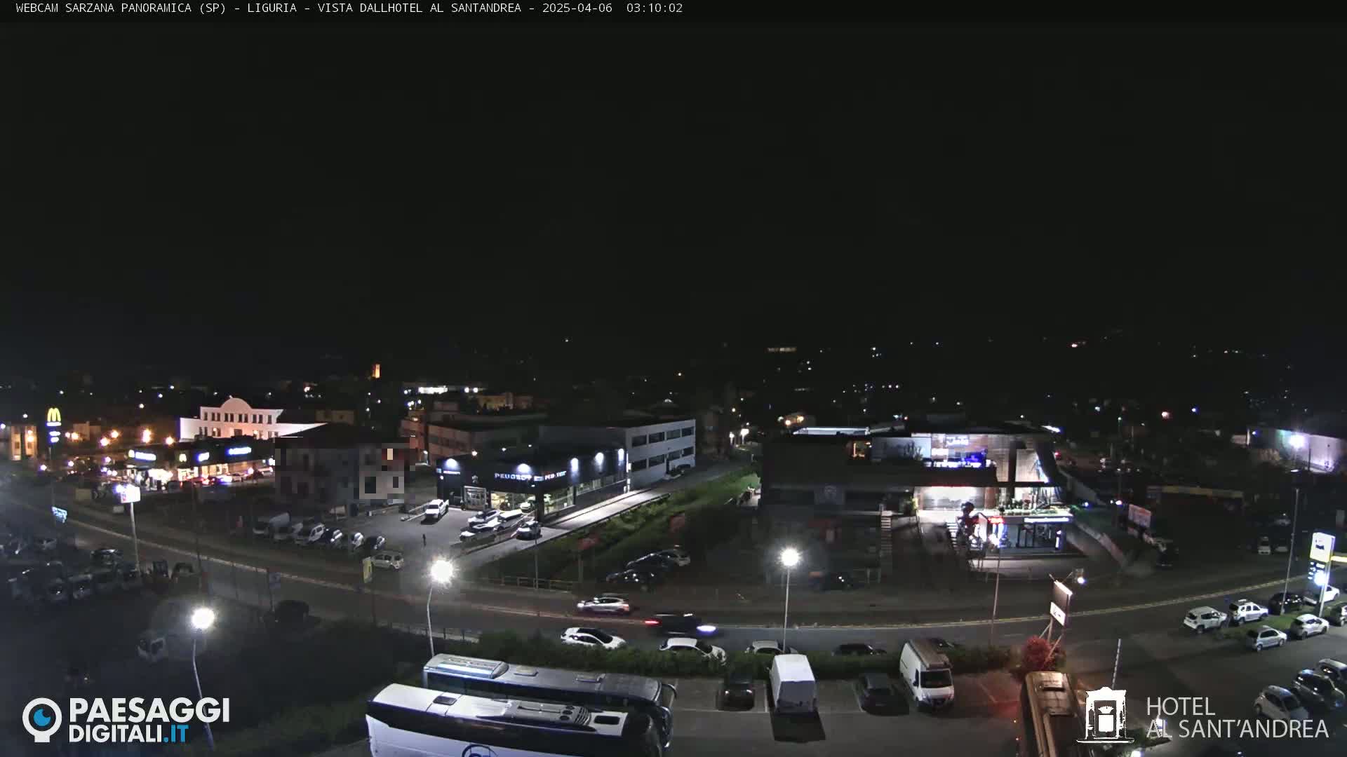 A nighttime aerial view of a town's outskirts, showing roadways, buildings, and parked cars under a clear, dark sky.