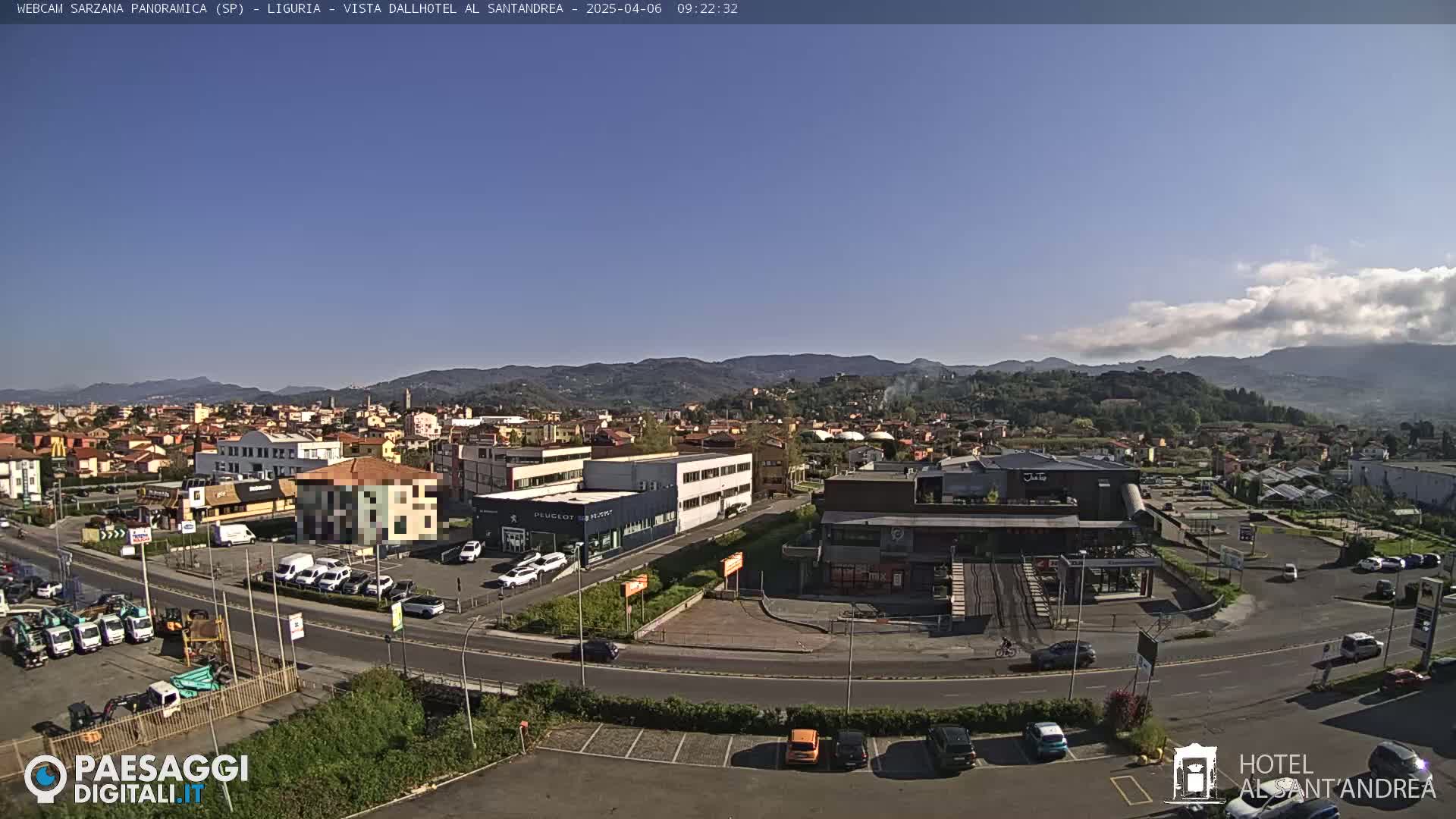 A clear, sunny day reveals a panoramic view of a town nestled in a valley, surrounded by rolling hills and mountains.