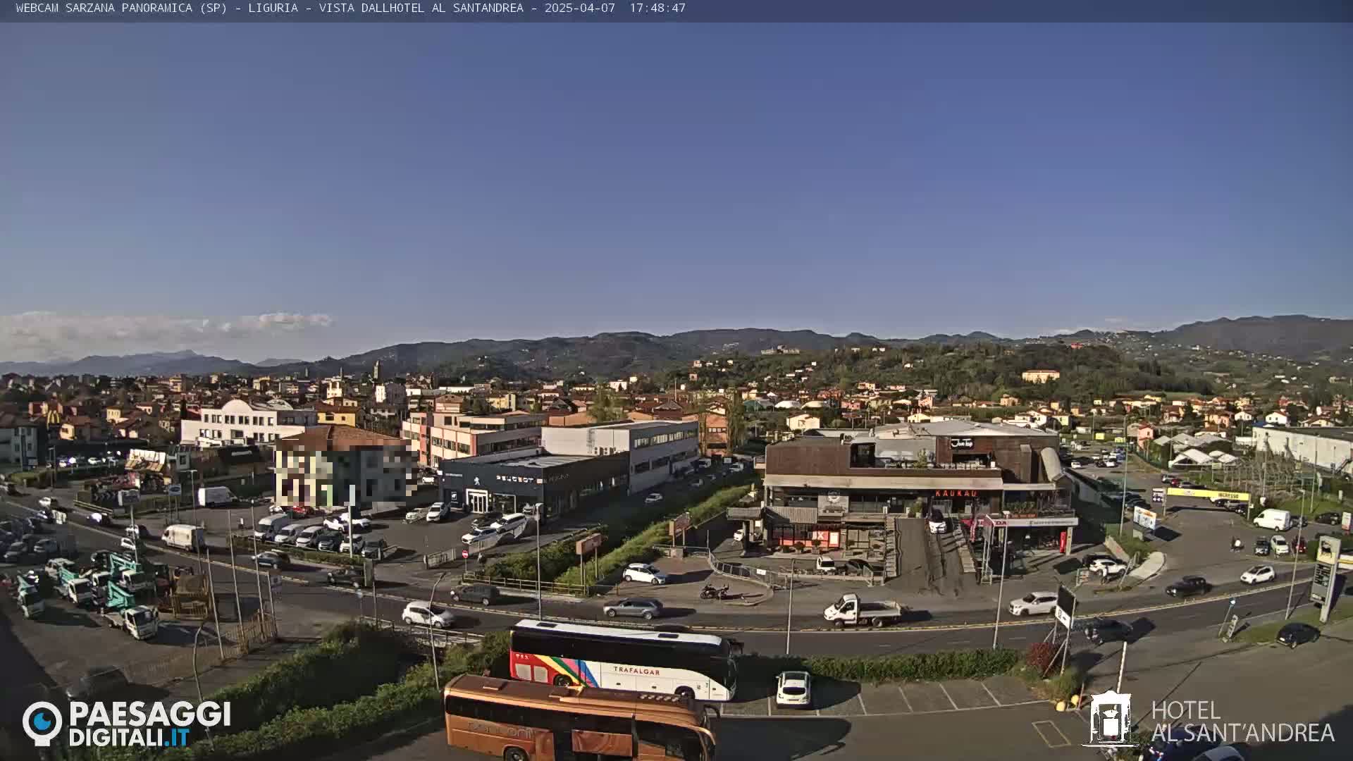 A panoramic view of a town on a clear, sunny day shows a mix of buildings, vehicles, and a large tour bus in a parking lot.