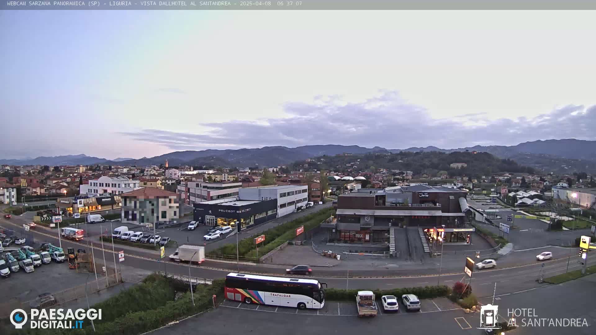 A long shot of a town under an overcast sky at dawn, showing several buildings, vehicles, and a bus in a parking lot.