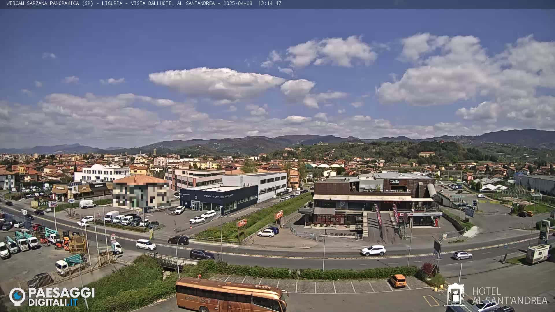 A partly sunny day overlooking a town with buildings, vehicles, and a large bus in a parking lot, mountains visible in the distance.
