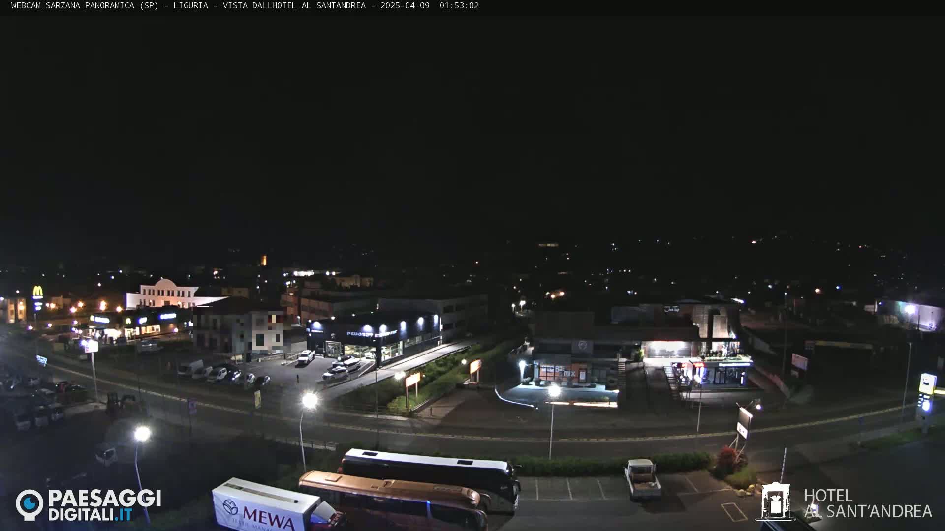 A nighttime aerial view of a town with a road curving through it, featuring several buildings, parked cars, and a couple of buses.