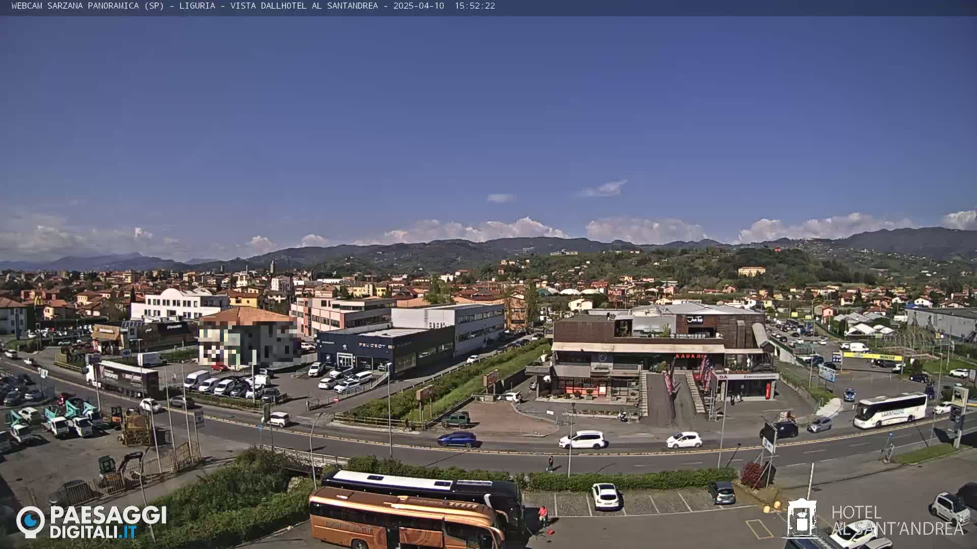 A sunny day reveals a panoramic view of a town nestled among rolling hills, with numerous cars, buses, and buildings visible, including a hotel.