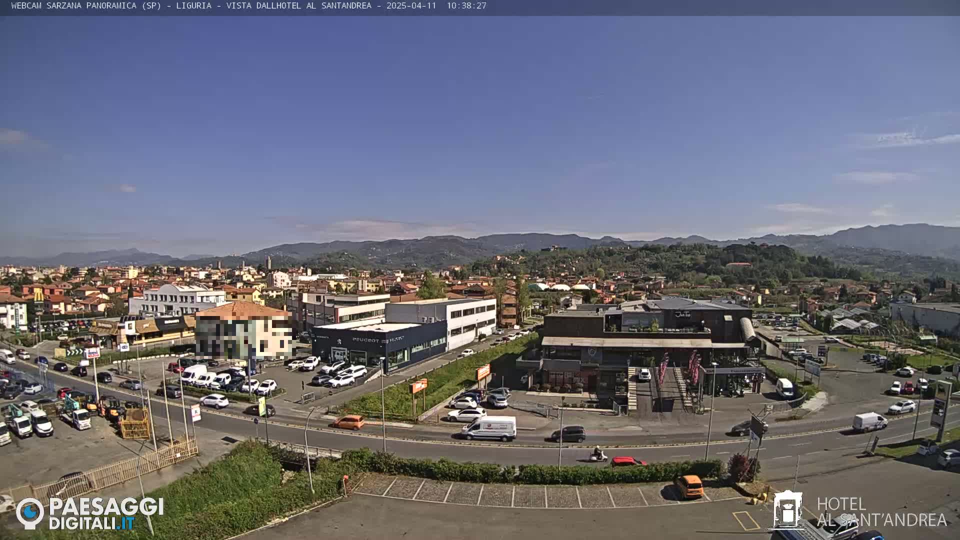 A sunny day reveals a sprawling town nestled in a valley, surrounded by hills and mountains, with numerous cars parked in lots surrounding buildings and businesses.