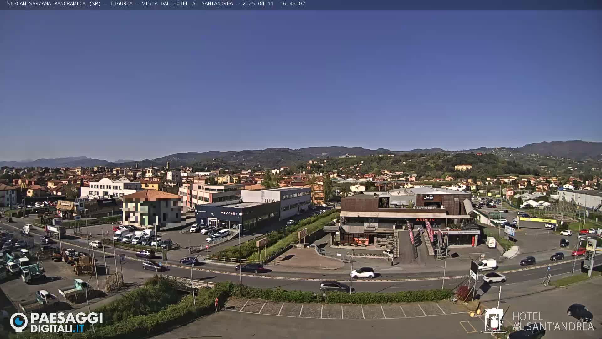 A sunny, clear day reveals a panoramic view of a town nestled among rolling hills, with a parking lot and various buildings visible in the foreground.