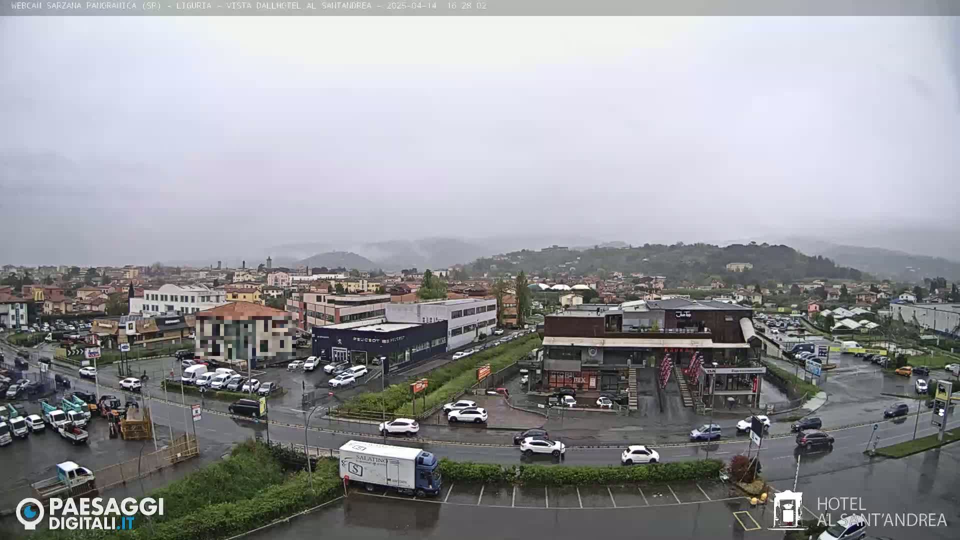 A rain-soaked town viewed from above, showing buildings, cars, and a truck in a parking lot, with hills in the background under an overcast sky.