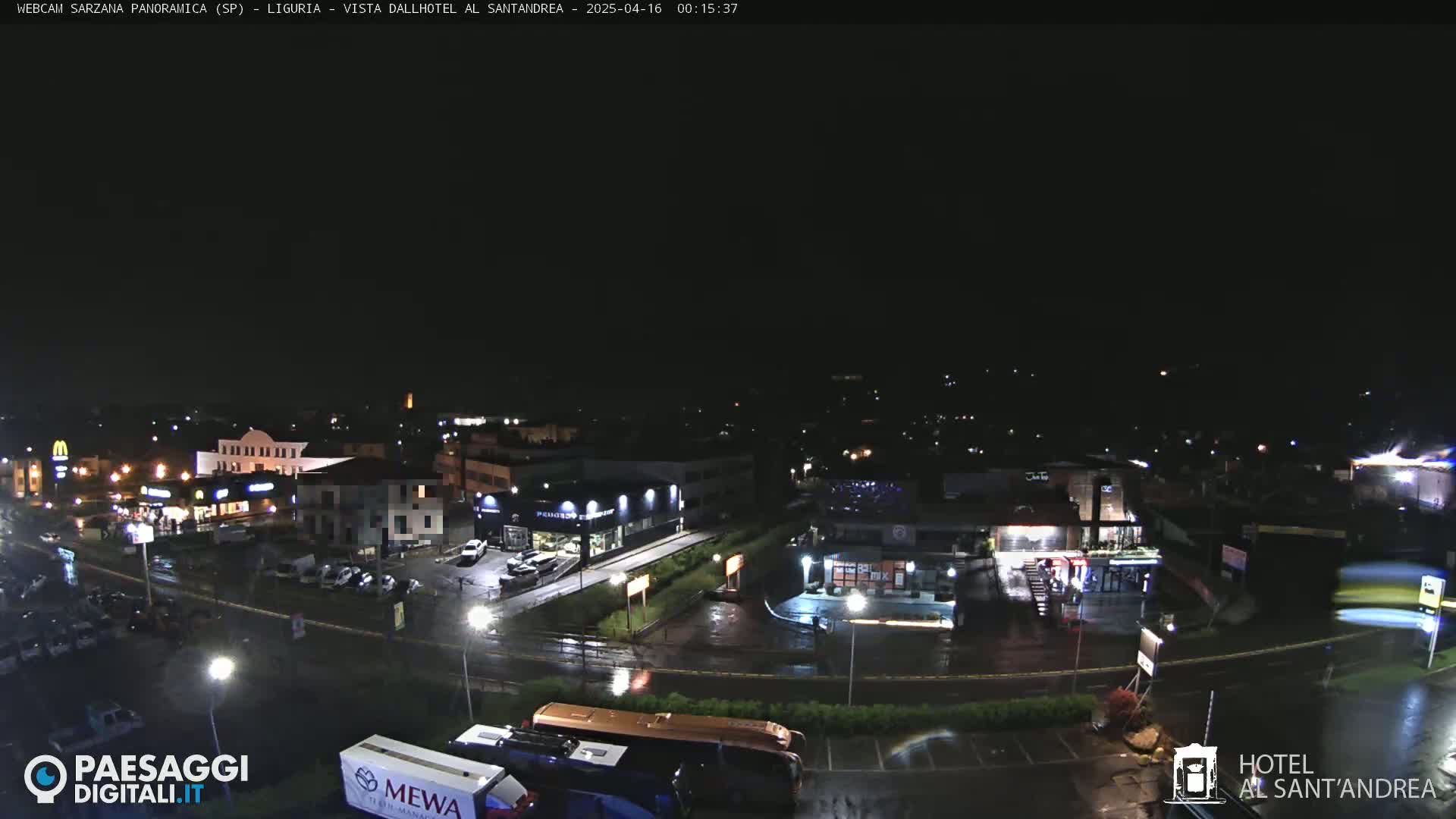 A nighttime view of a wet roadway and parking lot in front of several illuminated buildings, including restaurants and a hotel, under a dark sky.