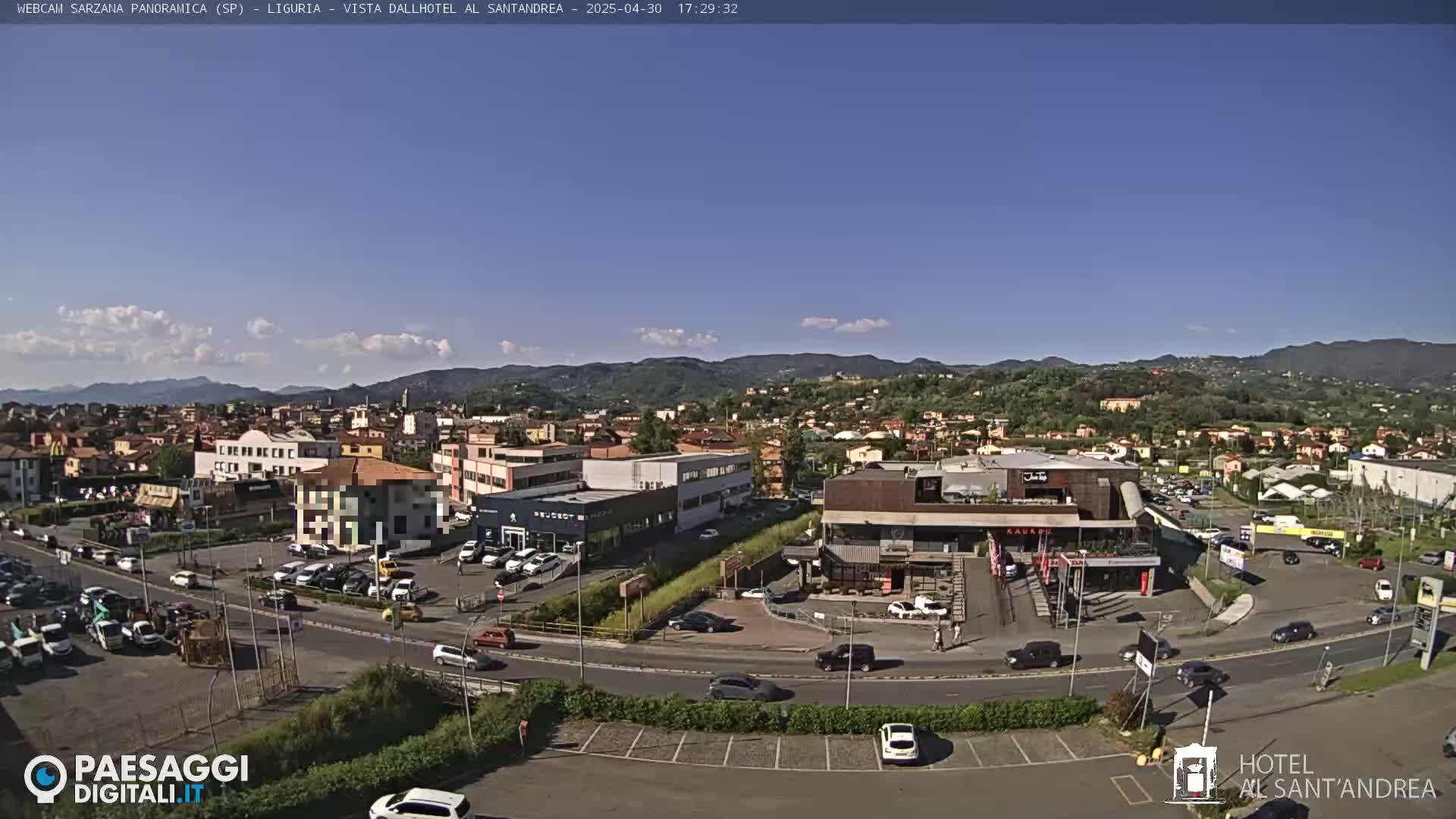 A sunny day reveals a panoramic view of a town nestled in a valley, with buildings, roads, and cars visible, surrounded by hills and mountains under a mostly clear sky.