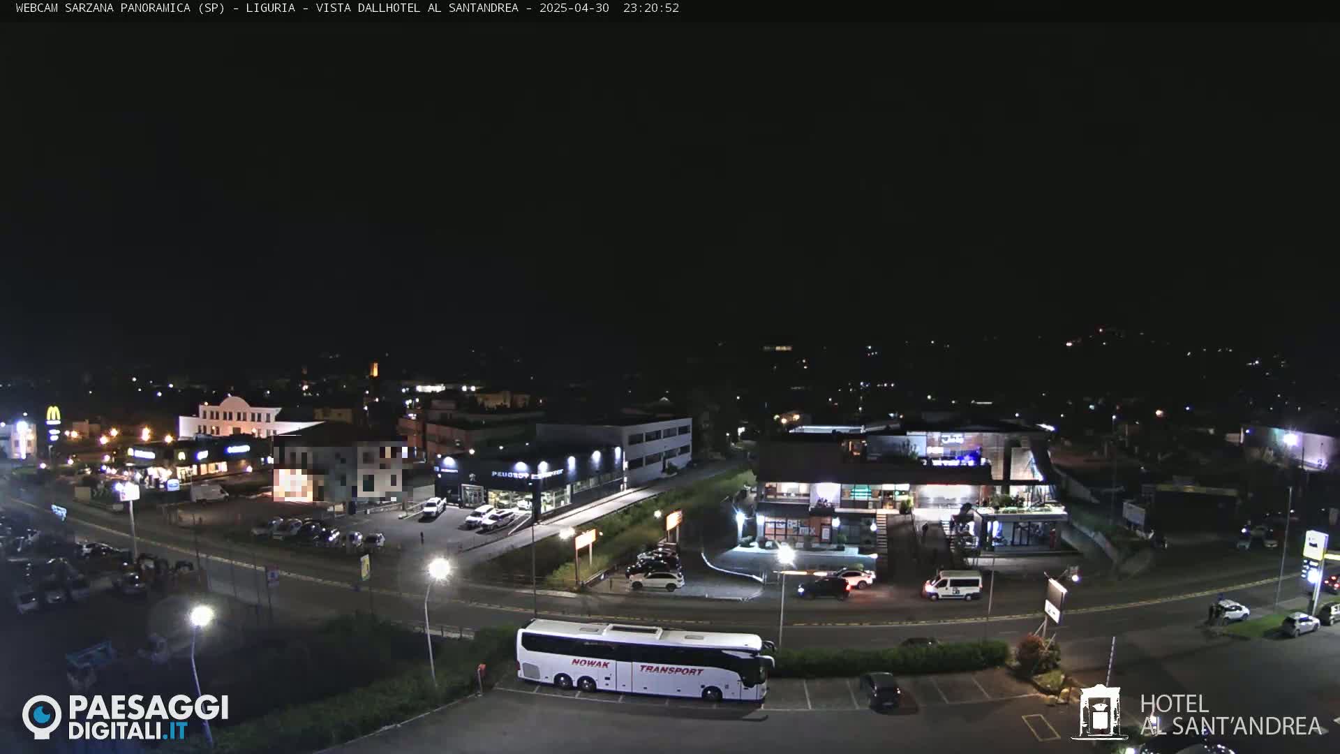 A nighttime, overhead view of a parking lot and roadway with various buildings, including a hotel and a bus, illuminated under artificial light.