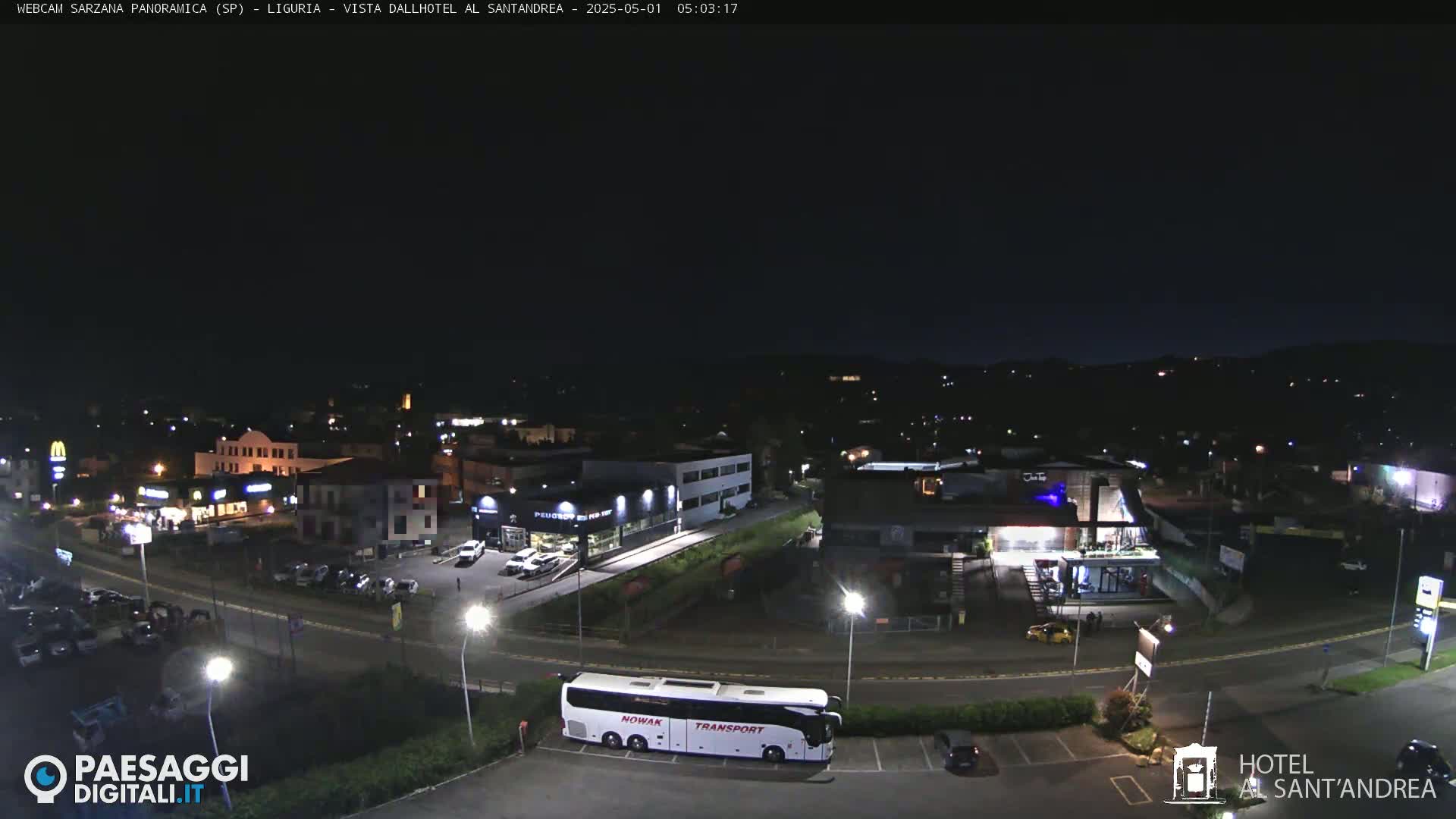 A nighttime, aerial view of a town with a bus parked in a parking lot, various buildings, and cars in the parking lot and on a road.