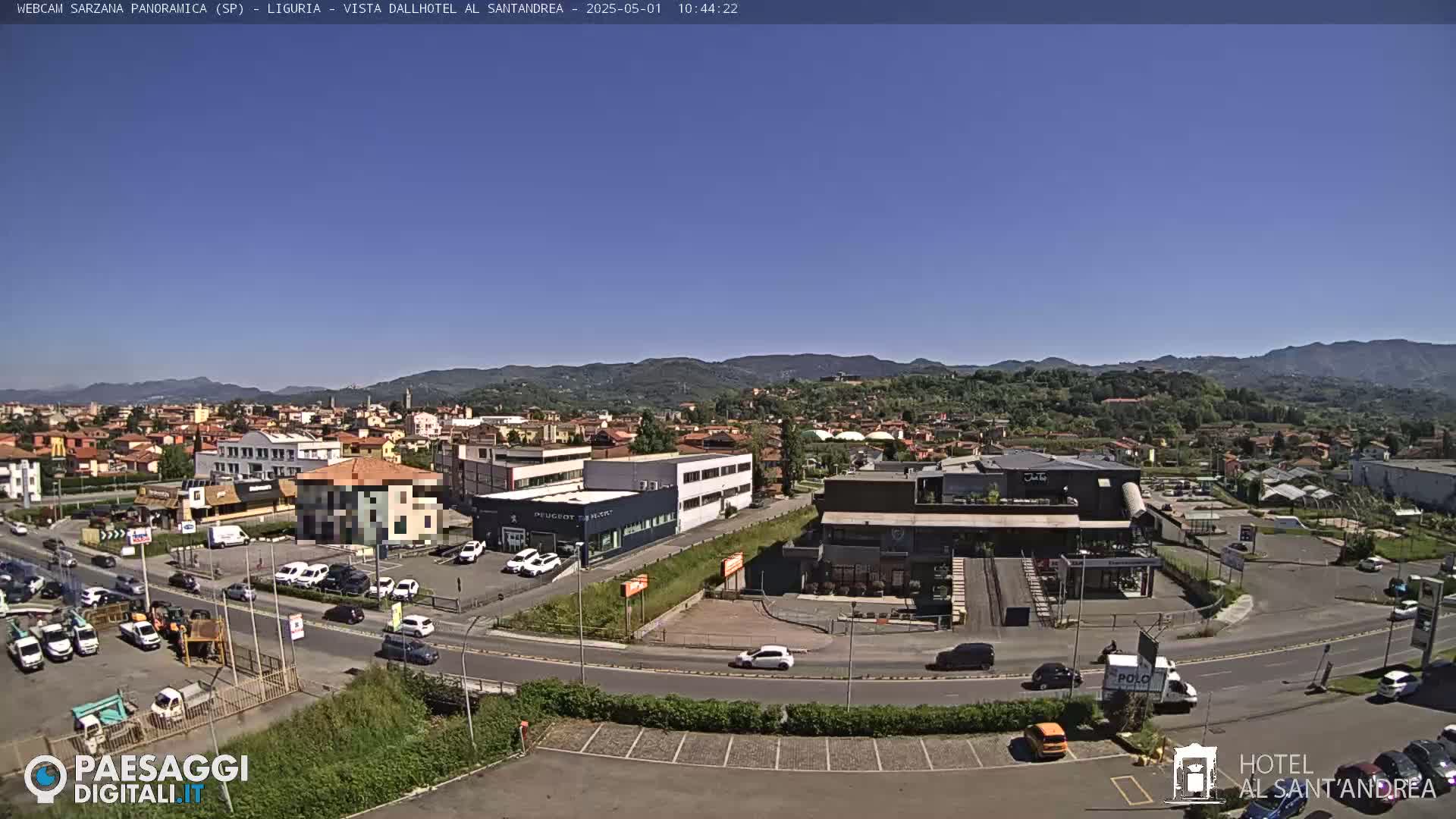 A sunny, clear day reveals a wide aerial view of a town, featuring buildings, roads, vehicles, and a surrounding landscape of hills under a vibrant blue sky.