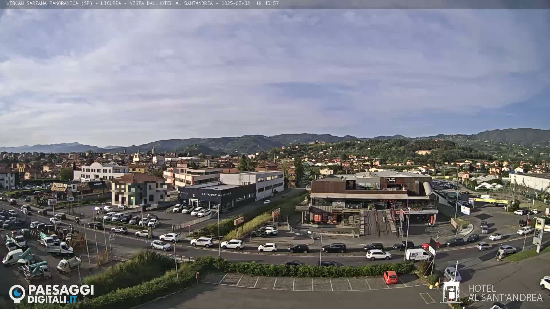 A panoramic view of a town on a partly cloudy day, showing a mix of buildings, vehicles in parking lots and on roadways, and a background of rolling hills.