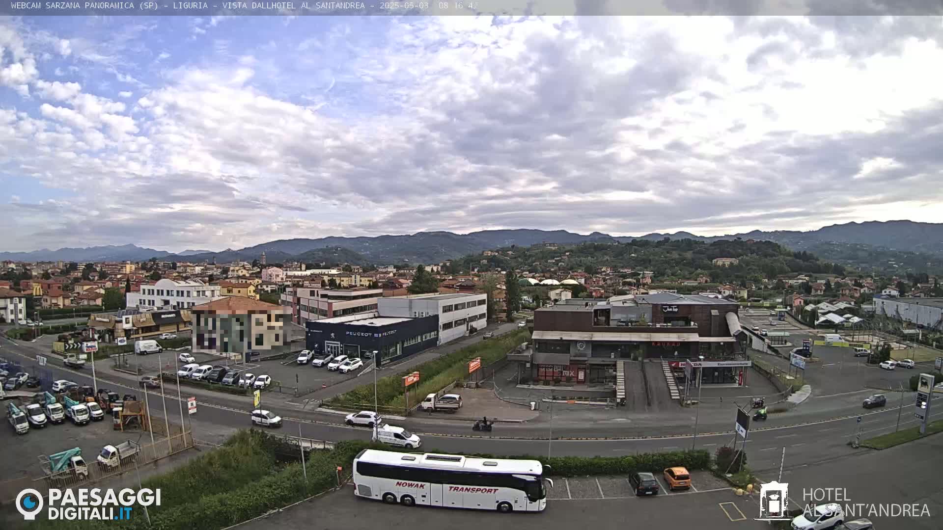 A mostly cloudy day reveals a panoramic view of a town nestled in a valley, with mountains in the distance, featuring a mix of buildings, vehicles, and a large tour bus in a parking lot.