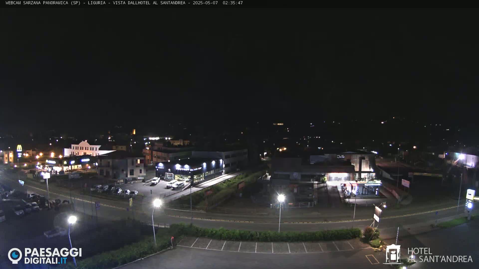 A nighttime aerial view of a town's outskirts, showing illuminated buildings, roads, and parking lots.