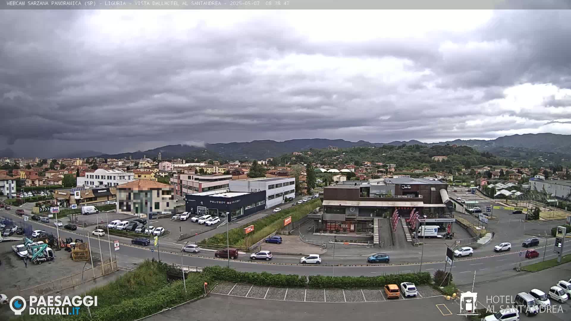 A panoramic view of a town nestled in a valley under a cloudy sky shows various buildings, including car dealerships and retail spaces, with cars parked in lots and driving on roads.
