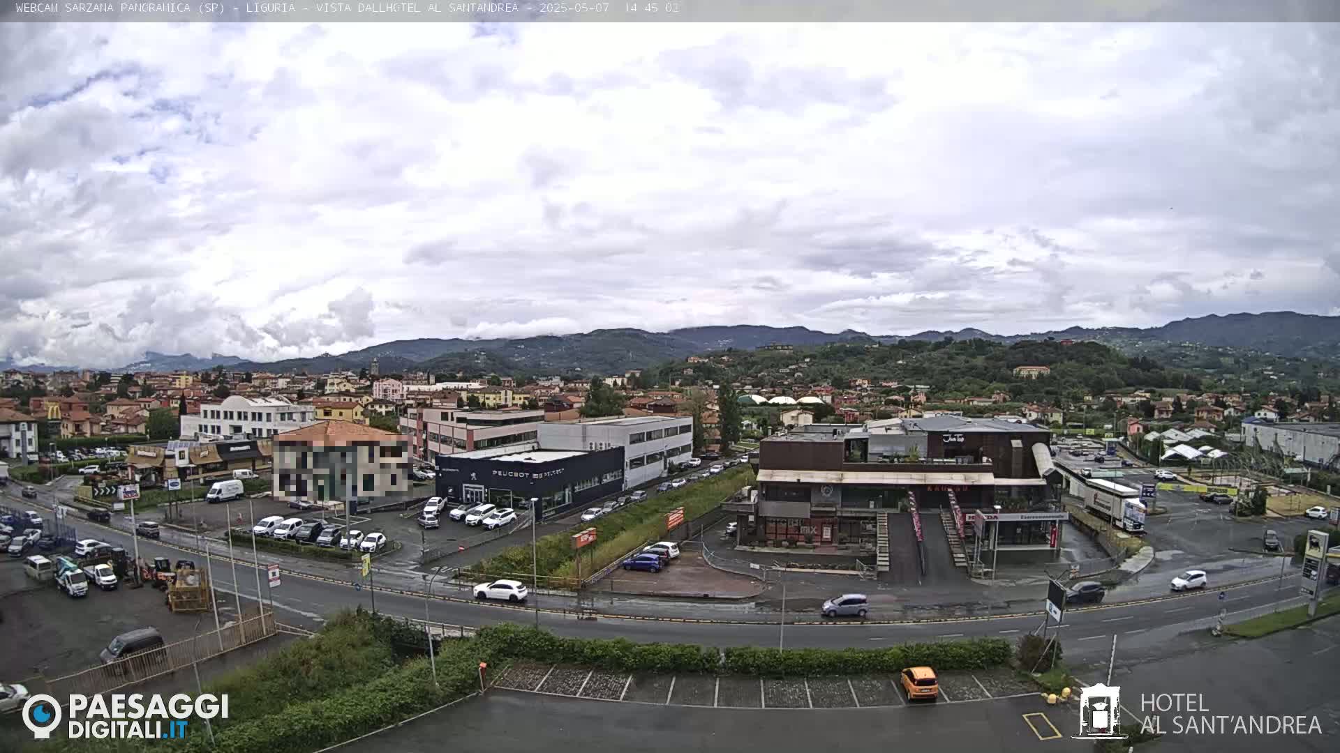 A partly cloudy day reveals a view of a town nestled among hills, with various buildings, cars, and a road visible in the foreground.