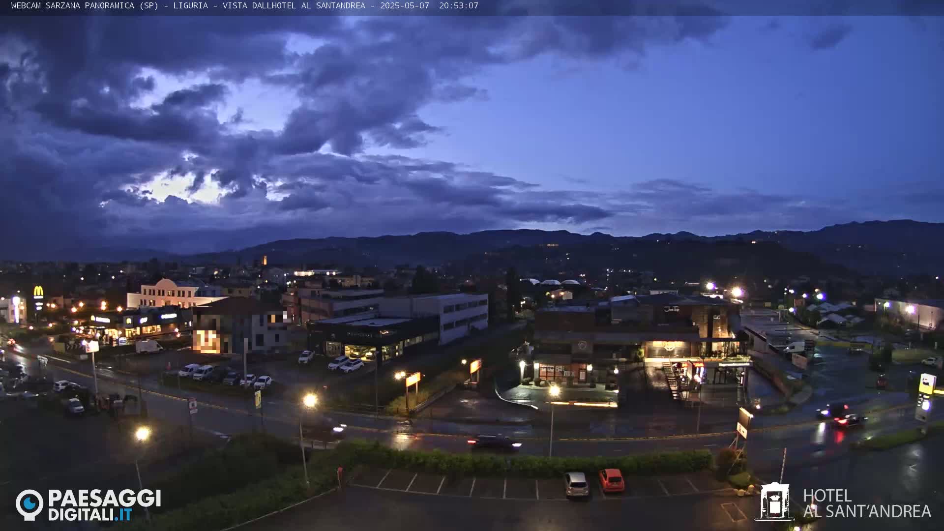 A nighttime view of a town with a partly cloudy sky, showing buildings, roads, cars, and mountains in the background under a dark blue sky.