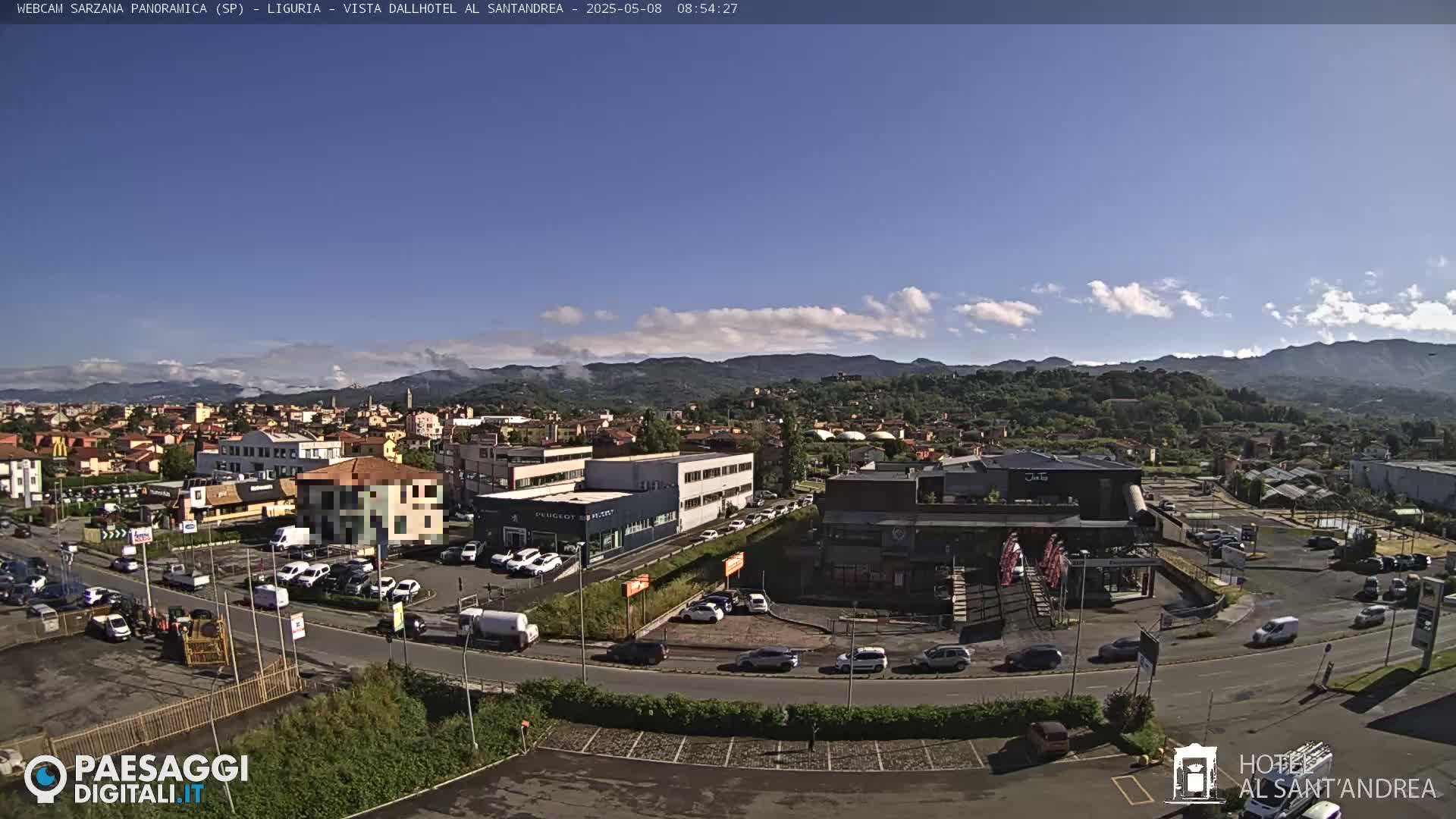 A panoramic view of a town nestled in a valley under a mostly clear, sunny sky, with mountains visible in the background and numerous cars parked in lots surrounding buildings.