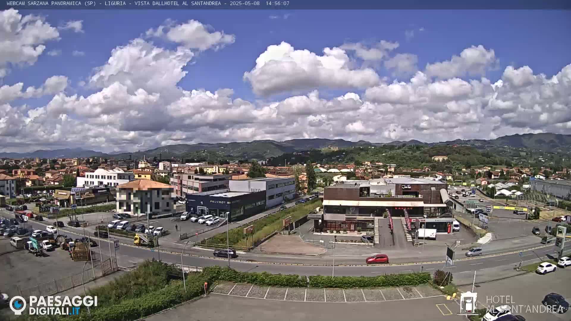 A partly cloudy day reveals a panoramic view of a town with buildings, roads, and parked cars, nestled against a backdrop of rolling hills.