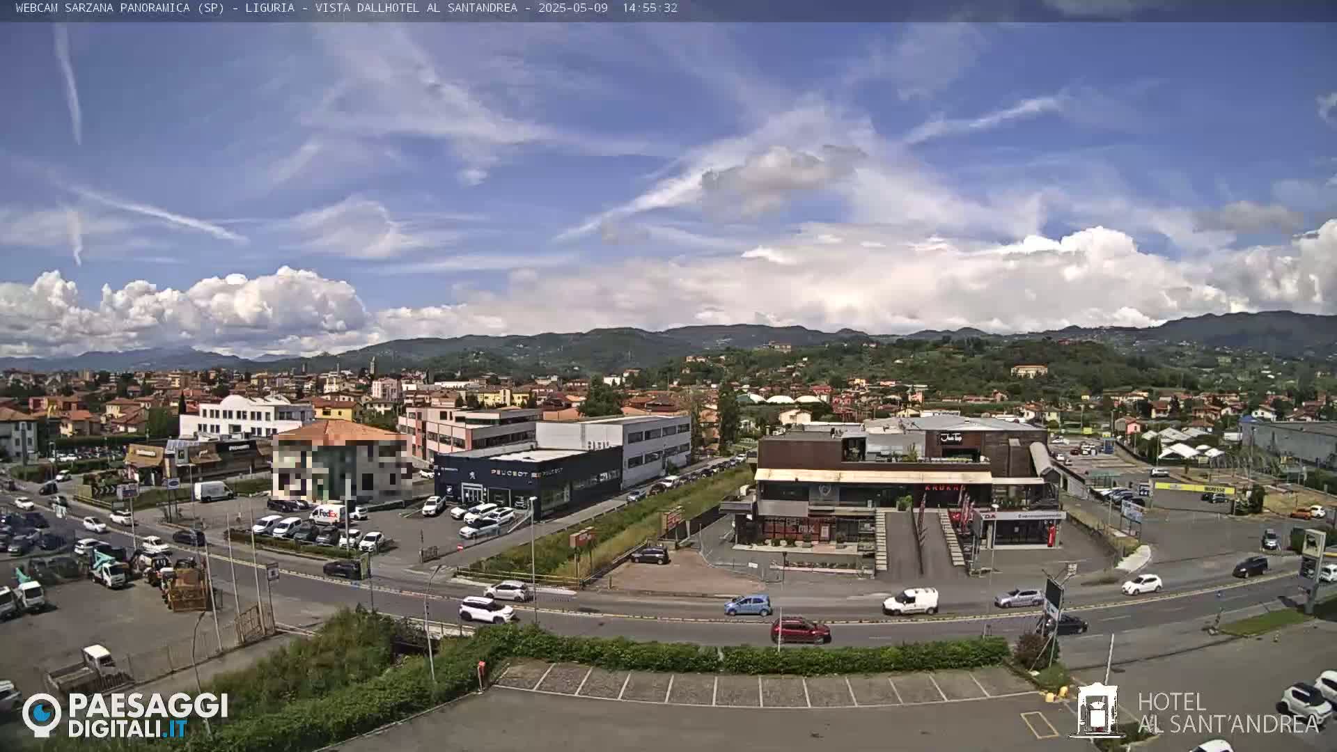 A partly cloudy day reveals a town nestled amongst green hills and mountains, with cars parked in lots surrounding buildings near a busy roadway.