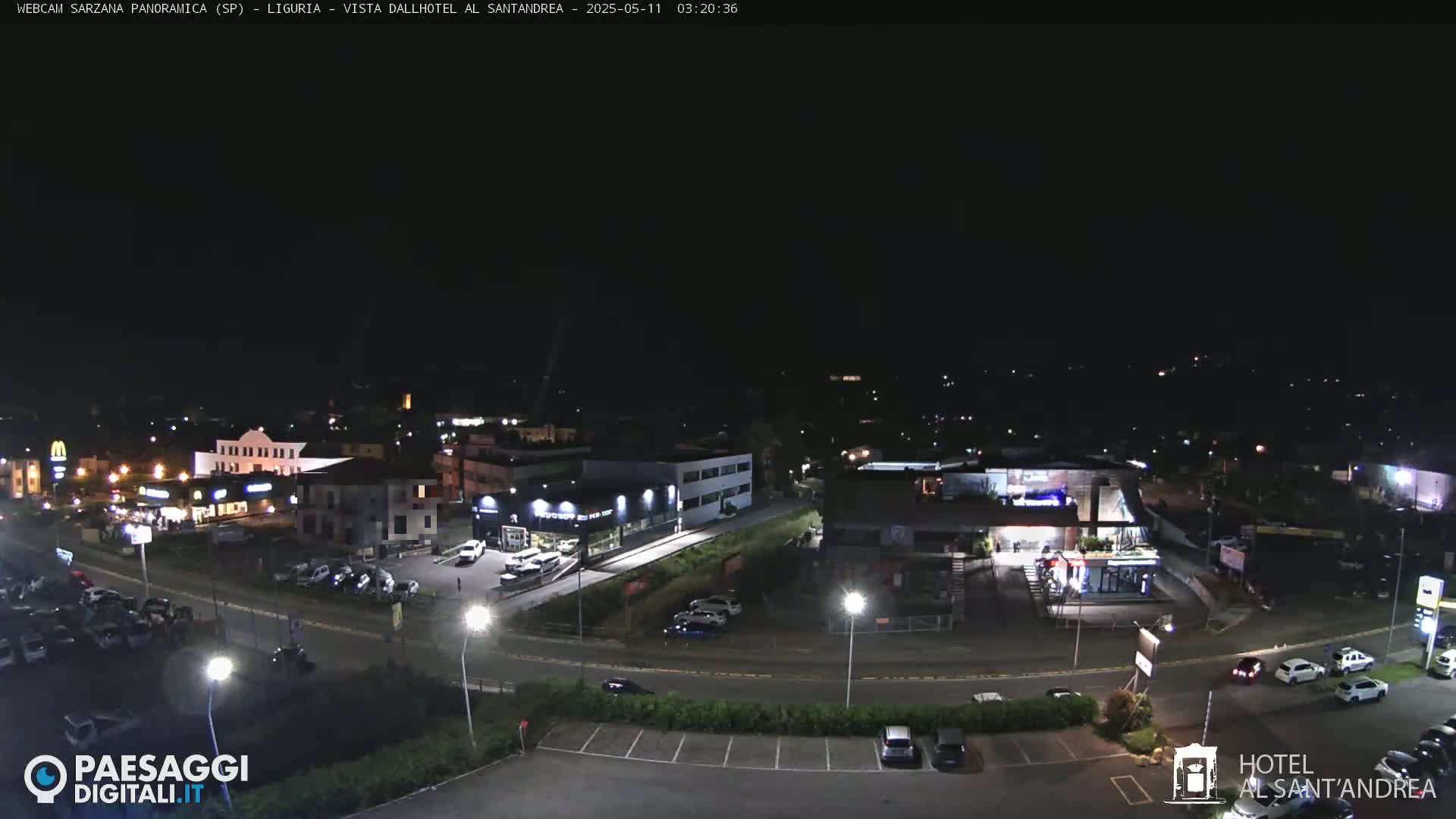 A nighttime, aerial view of a parking lot and buildings with scattered cars, illuminated by streetlights under a clear, dark sky.
