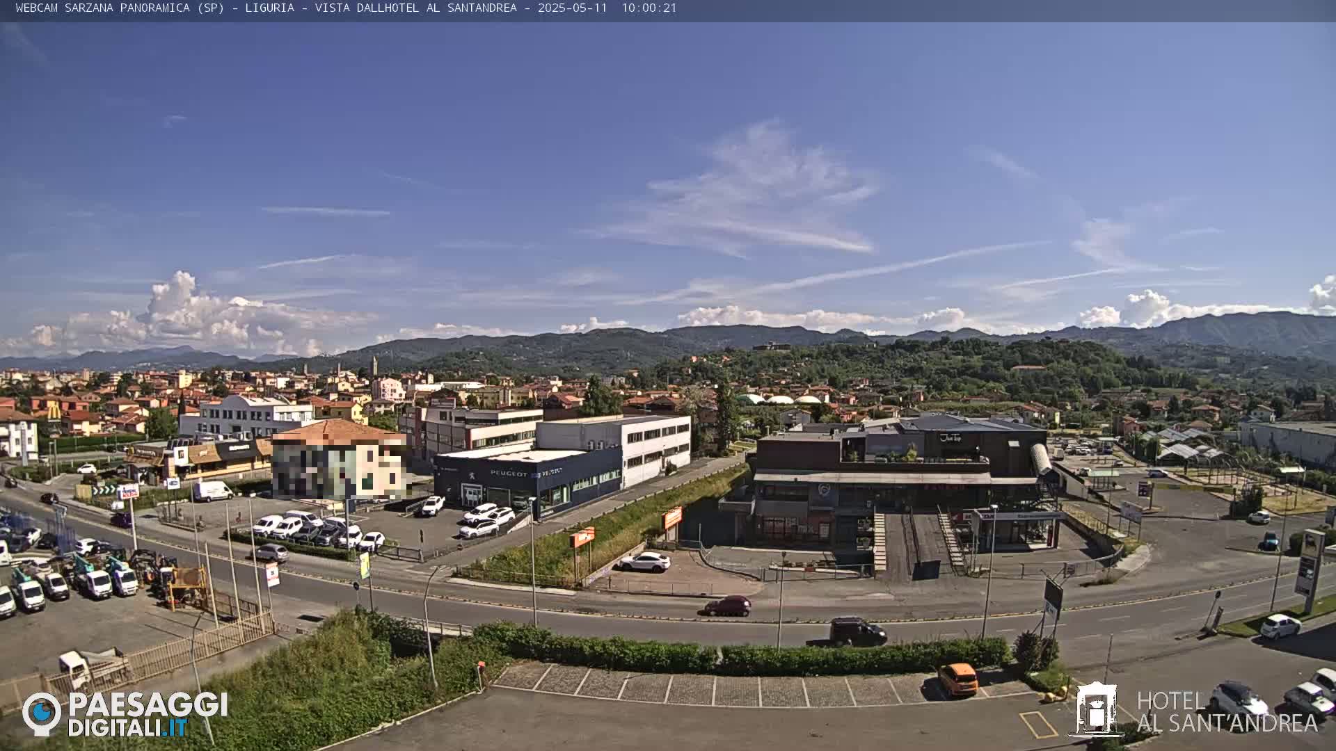 A sunny day reveals a sprawling town nestled in a valley, surrounded by verdant hills and a clear blue sky dotted with a few fluffy clouds.