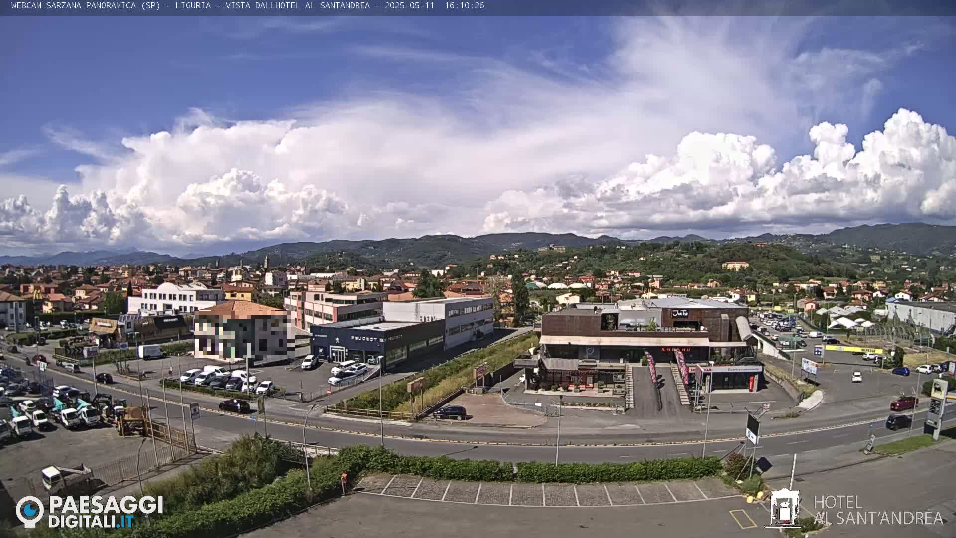 A partly cloudy day reveals a panoramic view of a town nestled among hills, with buildings, roads, and parked cars visible.