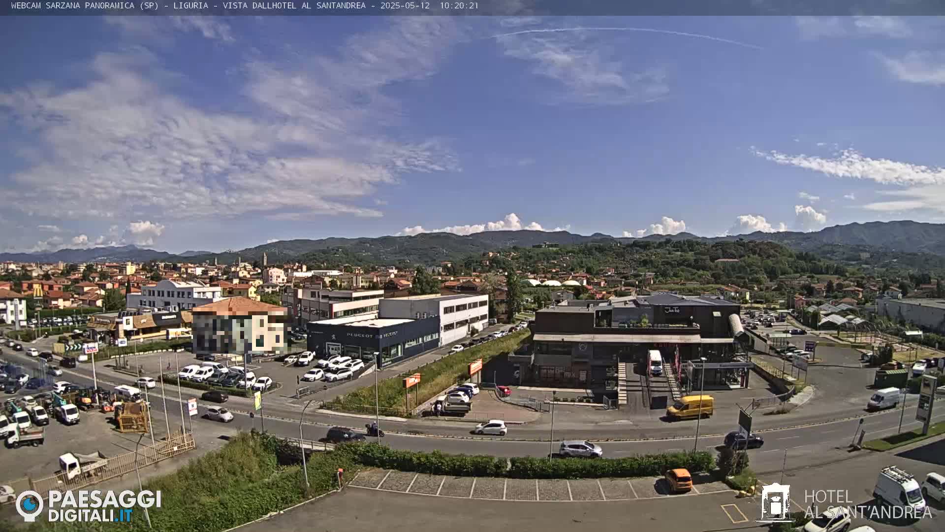 A sunny day reveals a panoramic view of a town, featuring buildings, roads with cars, and distant hills under a partly cloudy sky.