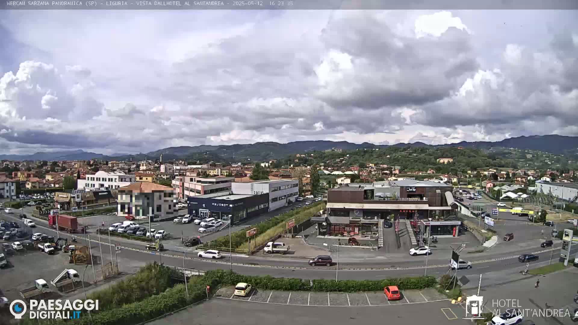 A partly cloudy day reveals a town nestled in a valley, with buildings, cars, and roads visible, mountains in the background.