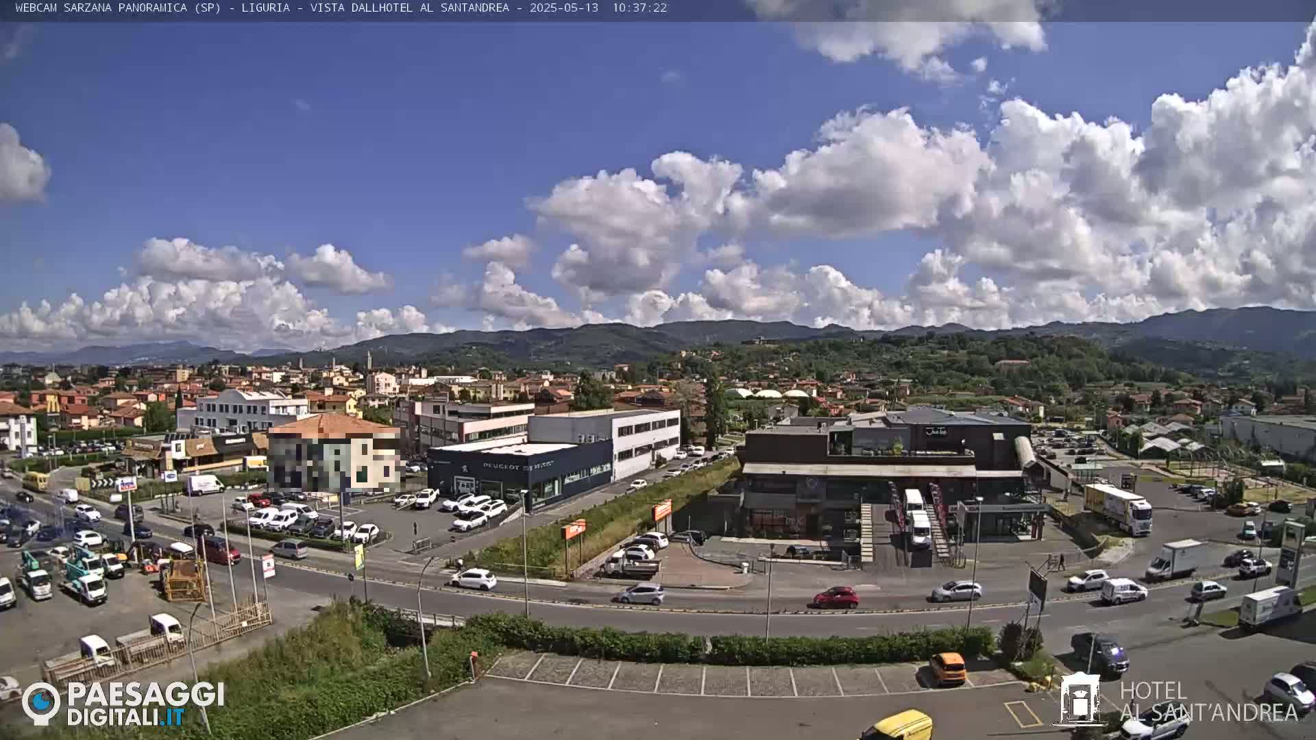A partly sunny day reveals a town nestled in a valley, with numerous buildings, cars, and trucks visible, and mountains in the distance.