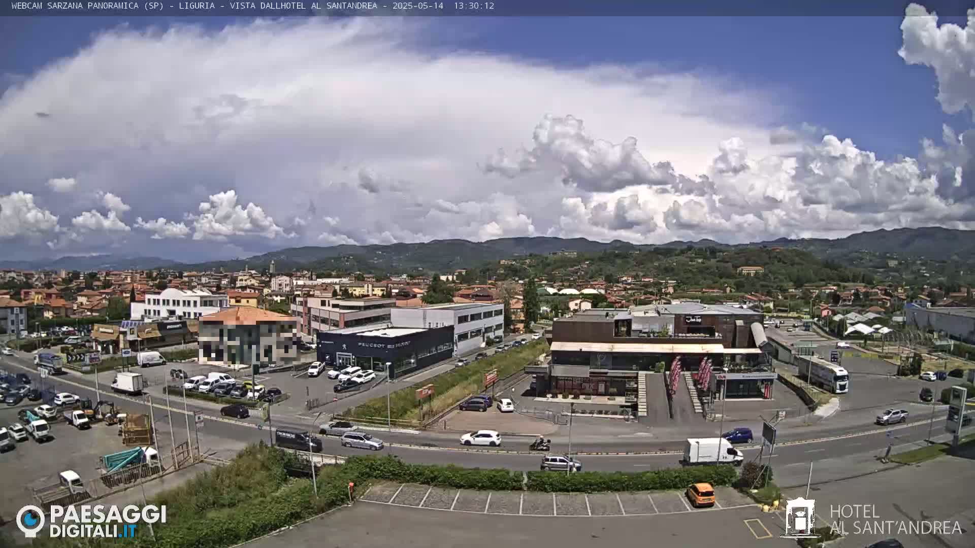 A partly cloudy day reveals a panoramic view of a town with buildings, roads, and parked cars, nestled against a backdrop of rolling hills.