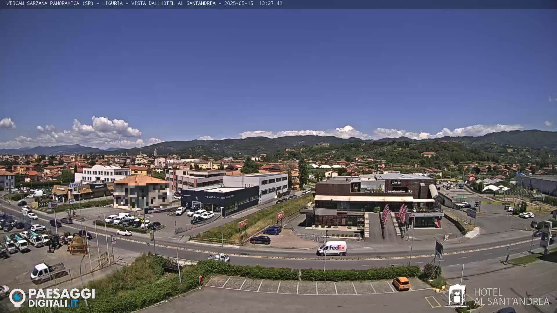 A sunny day reveals a panoramic view of a town nestled in a valley, surrounded by mountains under a partly cloudy sky.