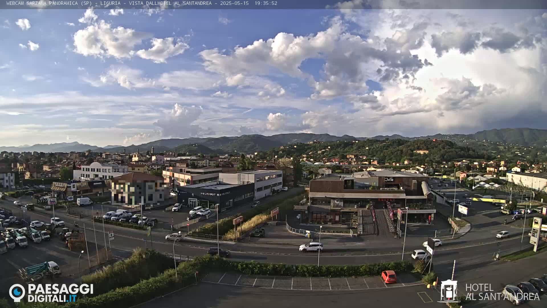A partly cloudy afternoon view of a town nestled in a valley, with mountains in the background and cars parked in lots near buildings along a road.