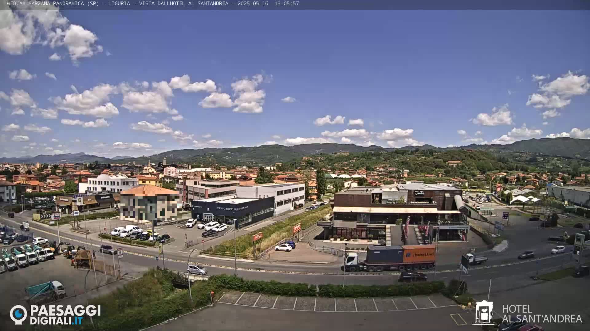 A sunny day with scattered clouds over a town nestled in a valley, with mountains in the background and various buildings, roads, and vehicles visible.