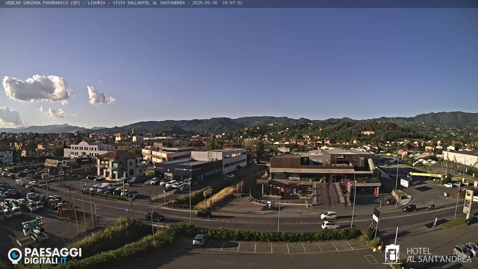A mostly sunny, clear day overlooks a town with buildings, roads, and cars, nestled amongst hills and mountains in the distance.