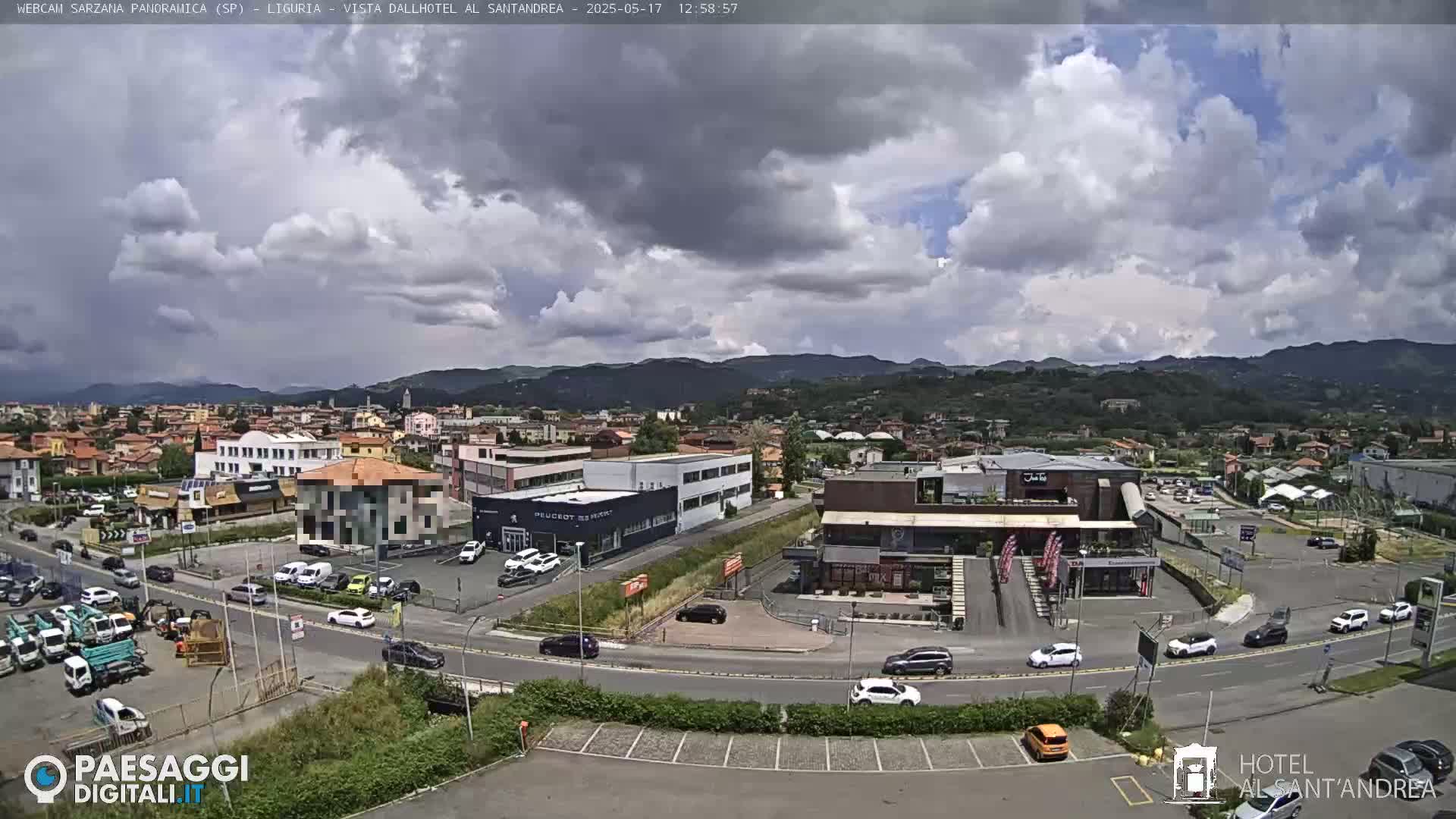 A partly cloudy day reveals a panoramic view of a town nestled in a valley, surrounded by hills, with cars parked in various lots and along roadways.